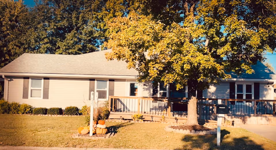A single-story care home with a wooden deck, pumpkins, and a large tree in the yard.
