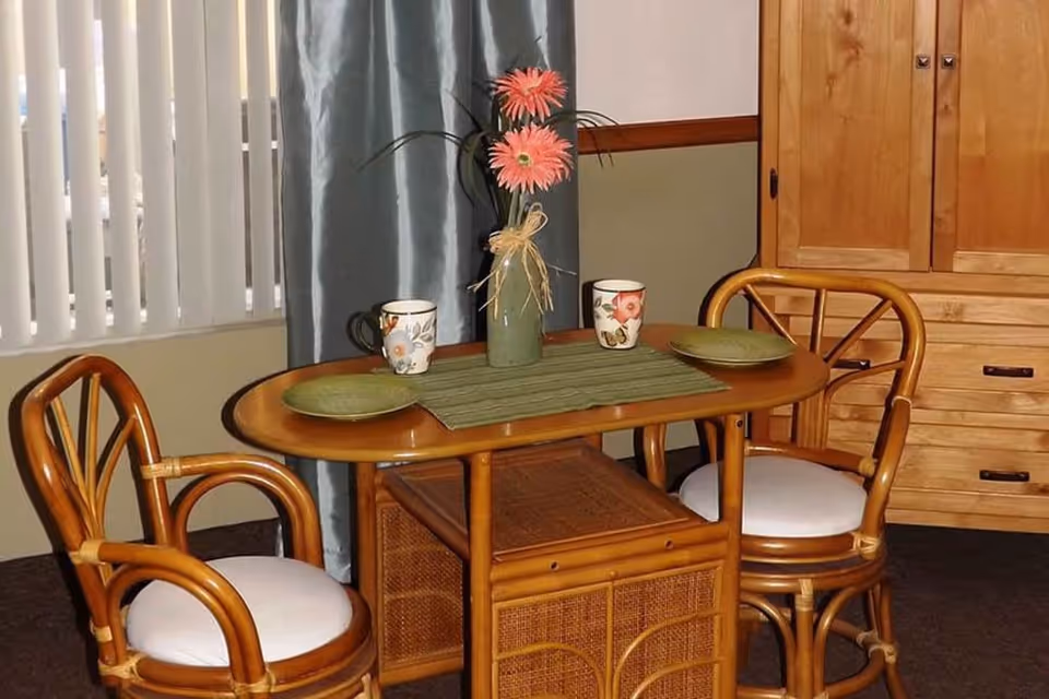 A small wooden dining table set with two chairs featuring white cushions. On the table are two floral-patterned mugs, two green plates, a green table runner, and a green vase with pink flowers. Behind the table is a window with vertical blinds and a blue curtain, and to the right is a wooden cabinet with drawers.