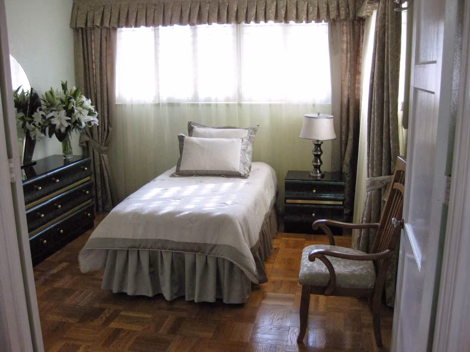 A neatly made single bed in a small bedroom with a bedside table and lamp, dresser with flowers, wooden chair, and a large curtained window.