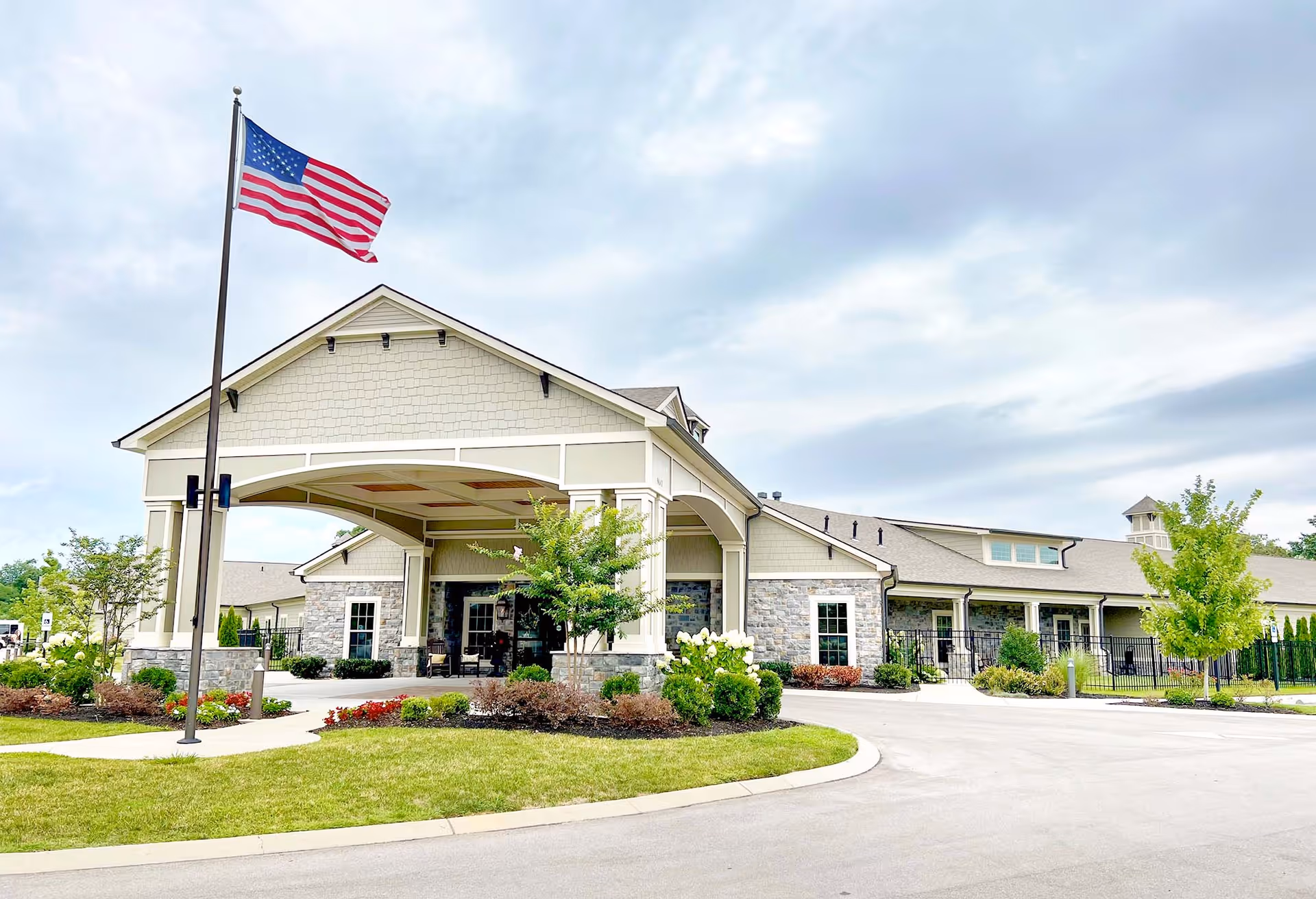 Front entrance of a single-story senior living facility with a covered porte-cochère, landscaped grounds, and an American flag flying.