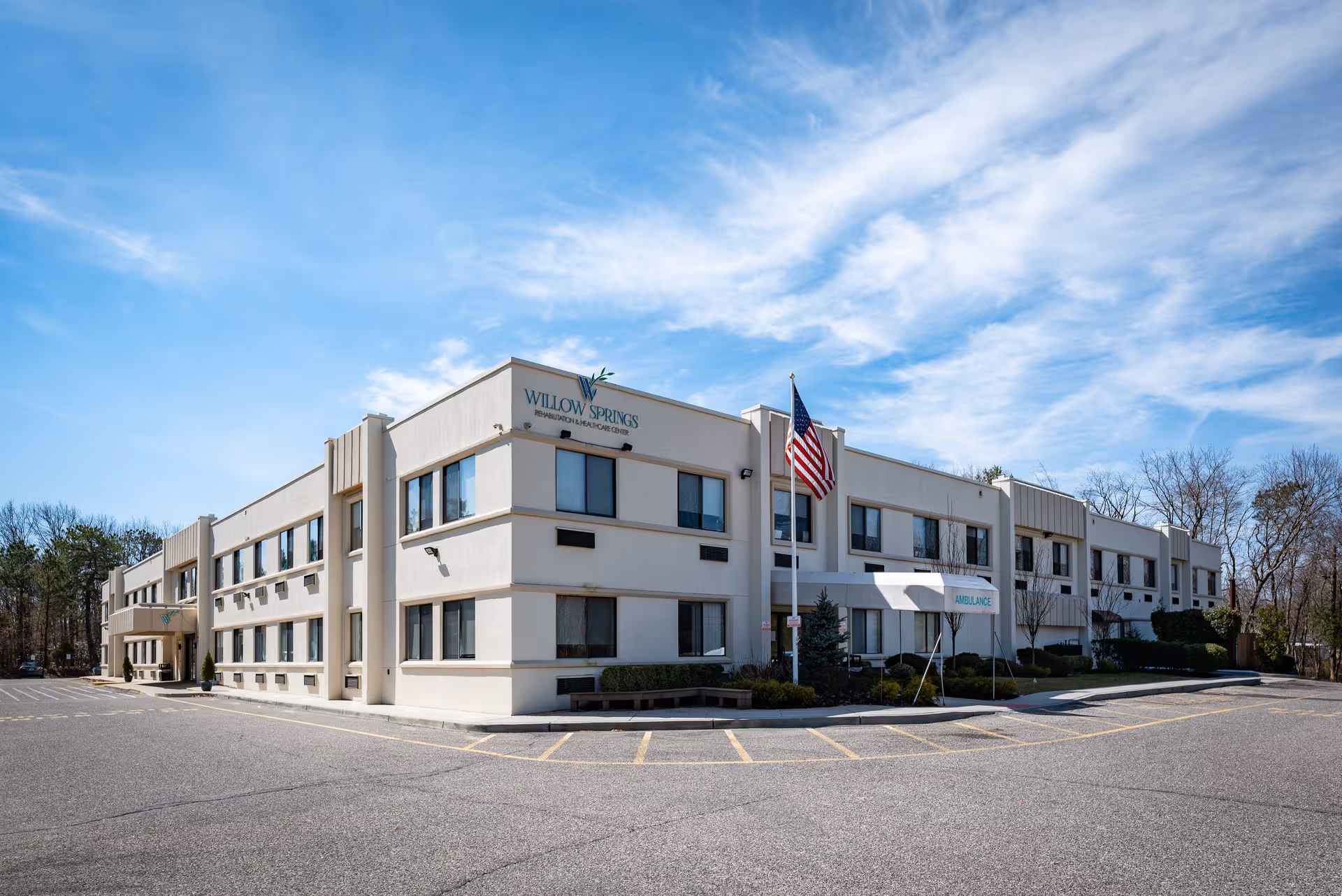 Two-story beige senior living building labeled Willow Springs with American flag and parking lot.