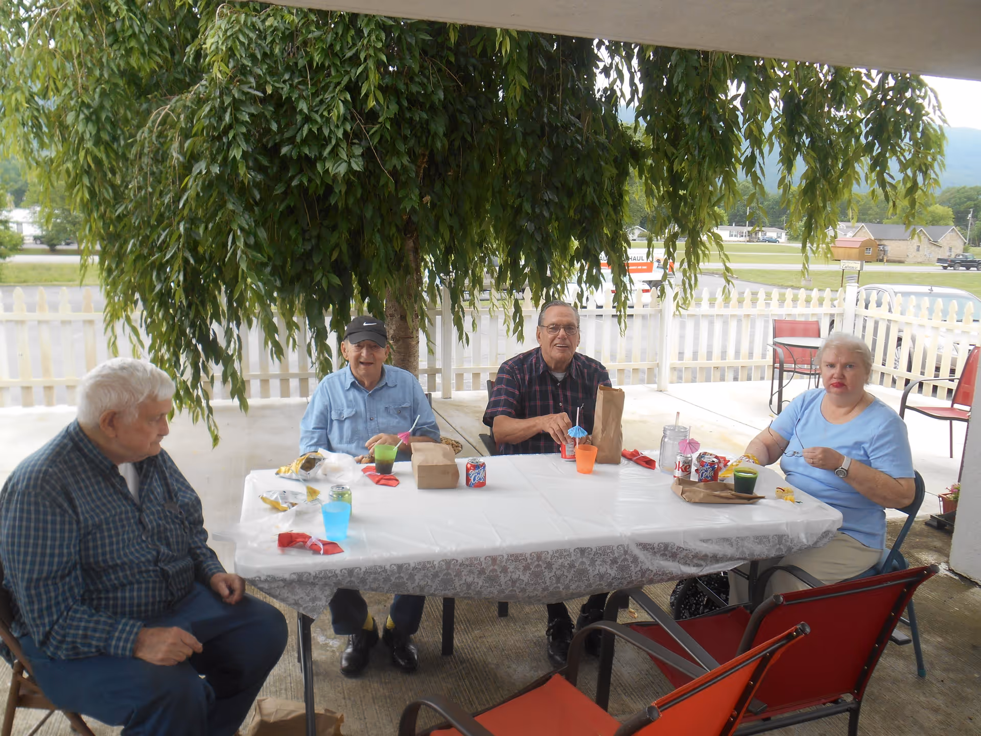 Four elderly people sitting around a rectangular table outdoors under a large leafy tree. They are having drinks and snacks, with a white picket fence and a road visible in the background.