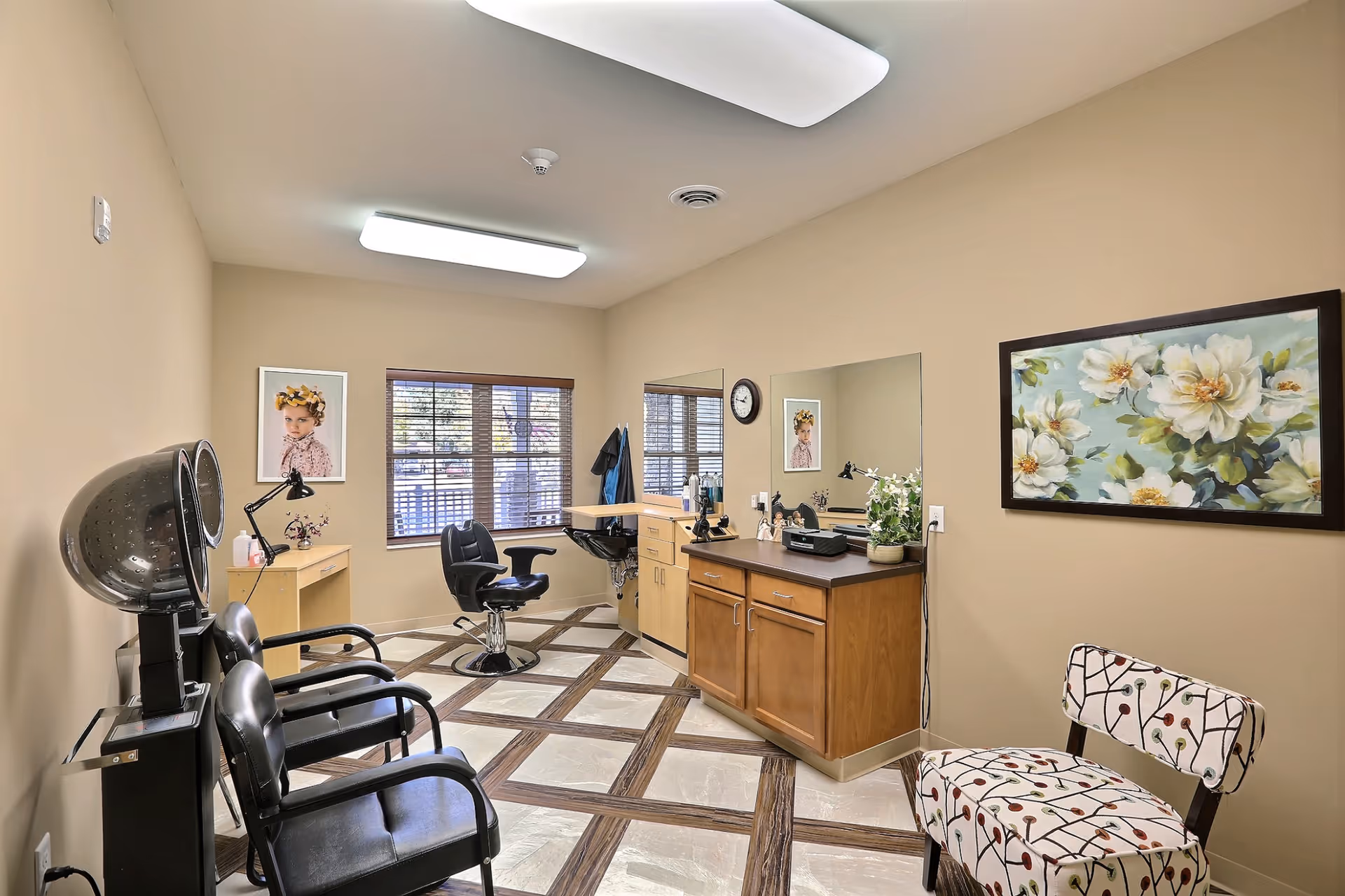 Interior view of a hair salon area in a senior living facility with two black salon chairs, a hair dryer, a styling station with a mirror, a small desk with a lamp, and a patterned chair. The room has beige walls, a large window with blinds, and decorative artwork on the walls.