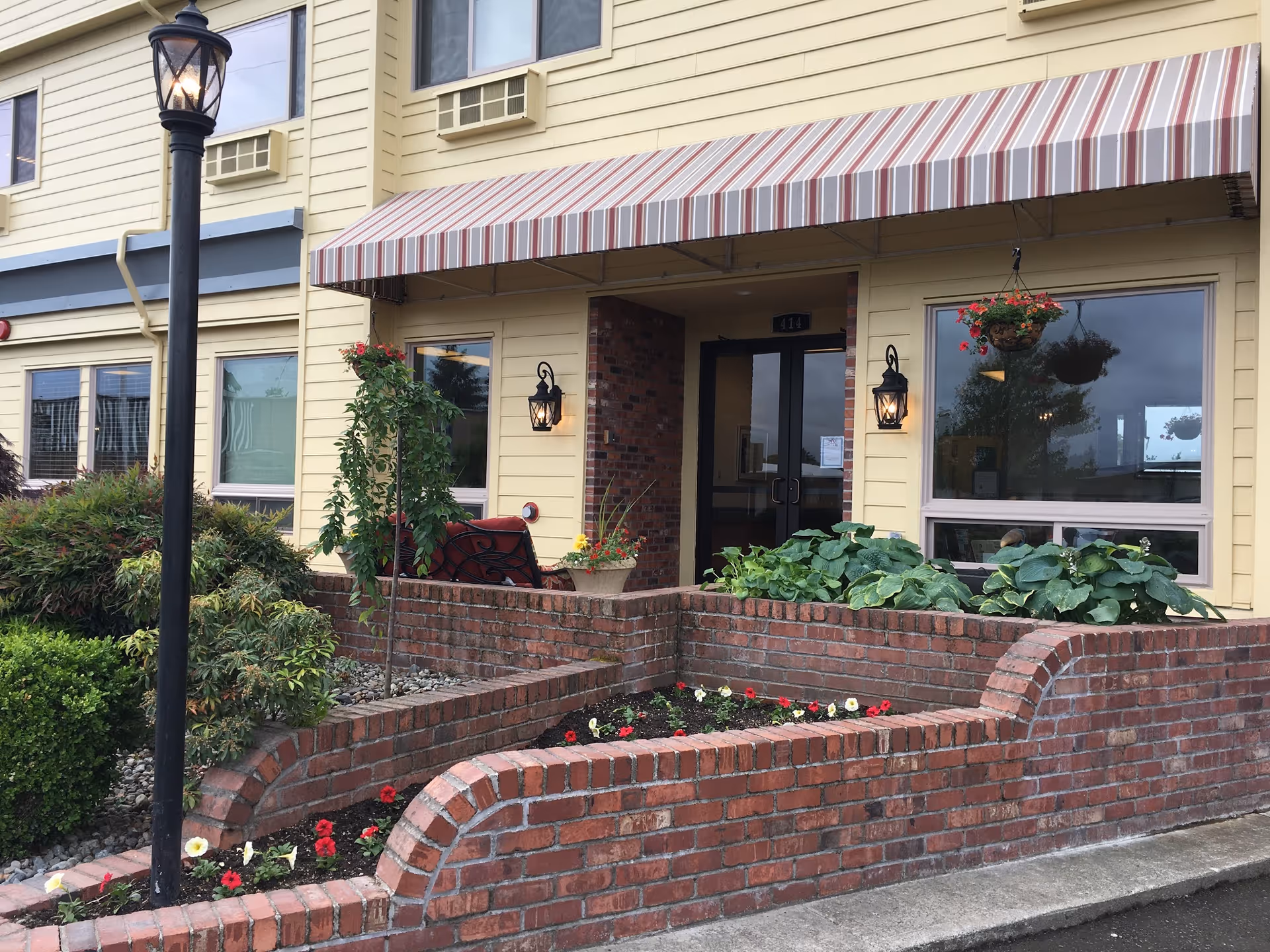 Exterior view of Auburn Post Acute facility showing a yellow building with a striped awning above the entrance. There are two black lantern-style wall lights on either side of the glass double doors. In front of the building is a brick planter with various green plants and flowers. A black street lamp is visible on the left side of the image.
