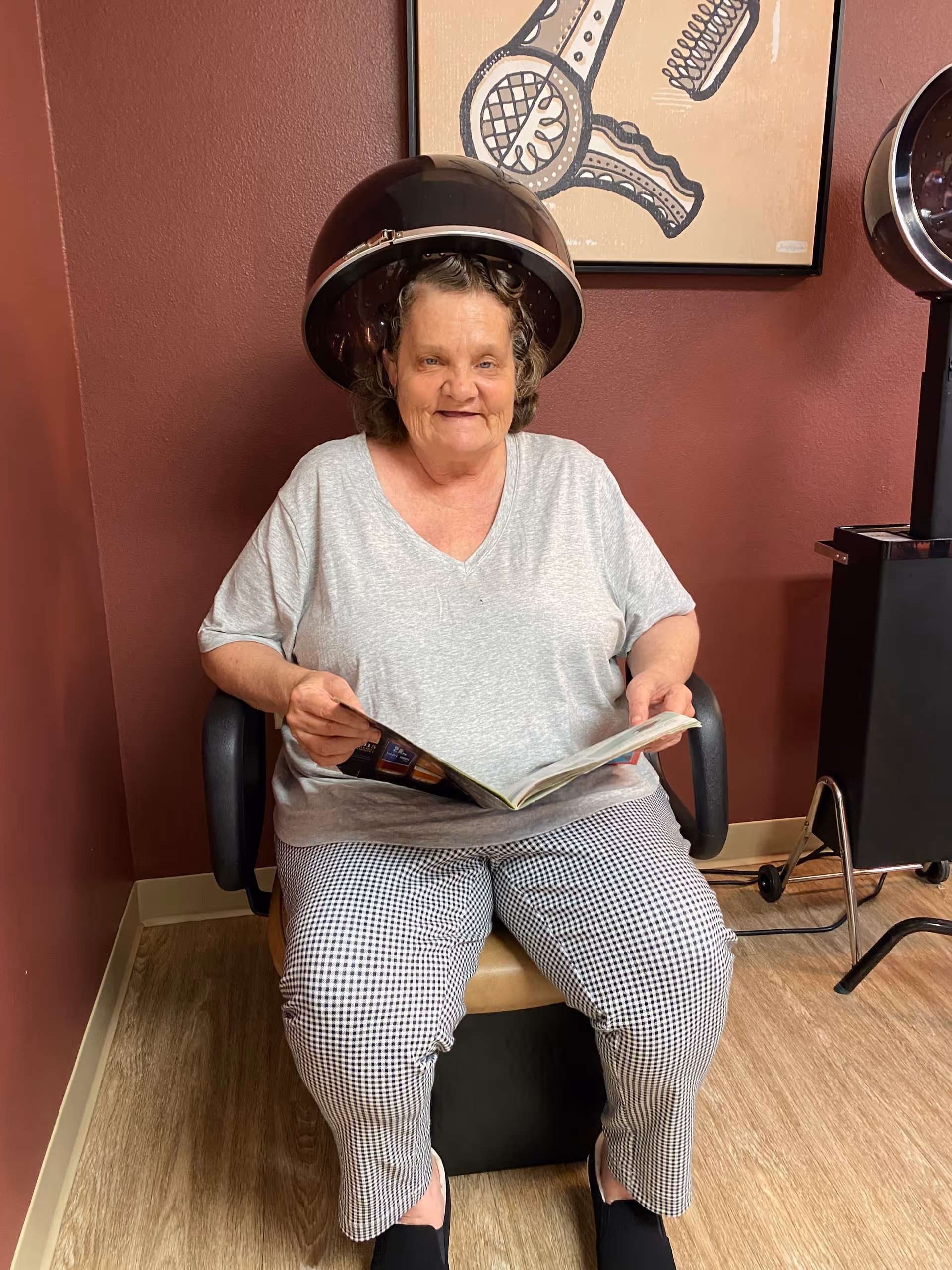 An elderly woman sitting under a hair dryer in a salon chair, reading a magazine. The room has maroon walls and wooden flooring, with a framed artwork of hair styling tools on the wall behind her.