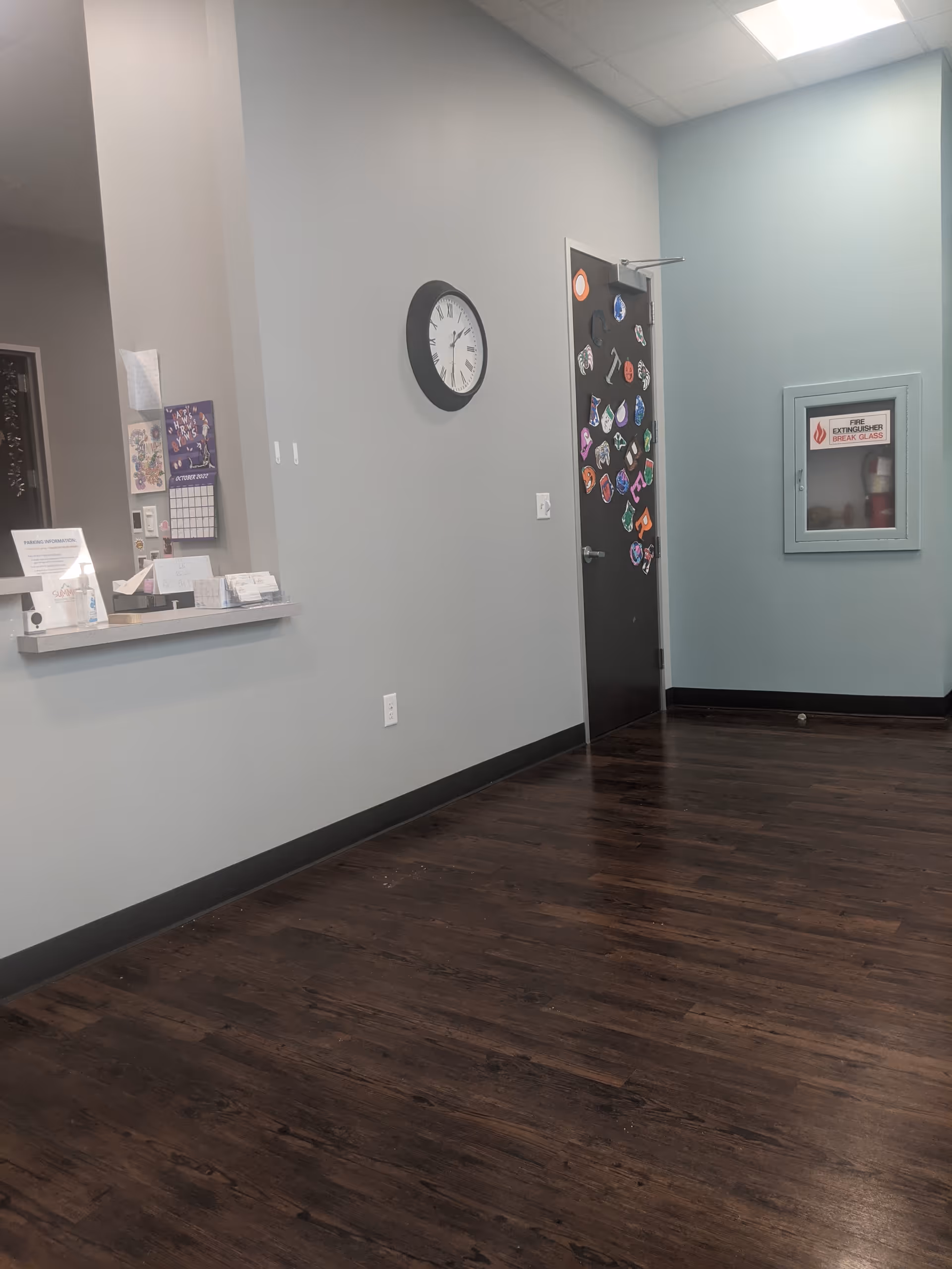 Indoor corridor with reception counter on left, clock on wall, magnet-covered door, and wooden flooring.