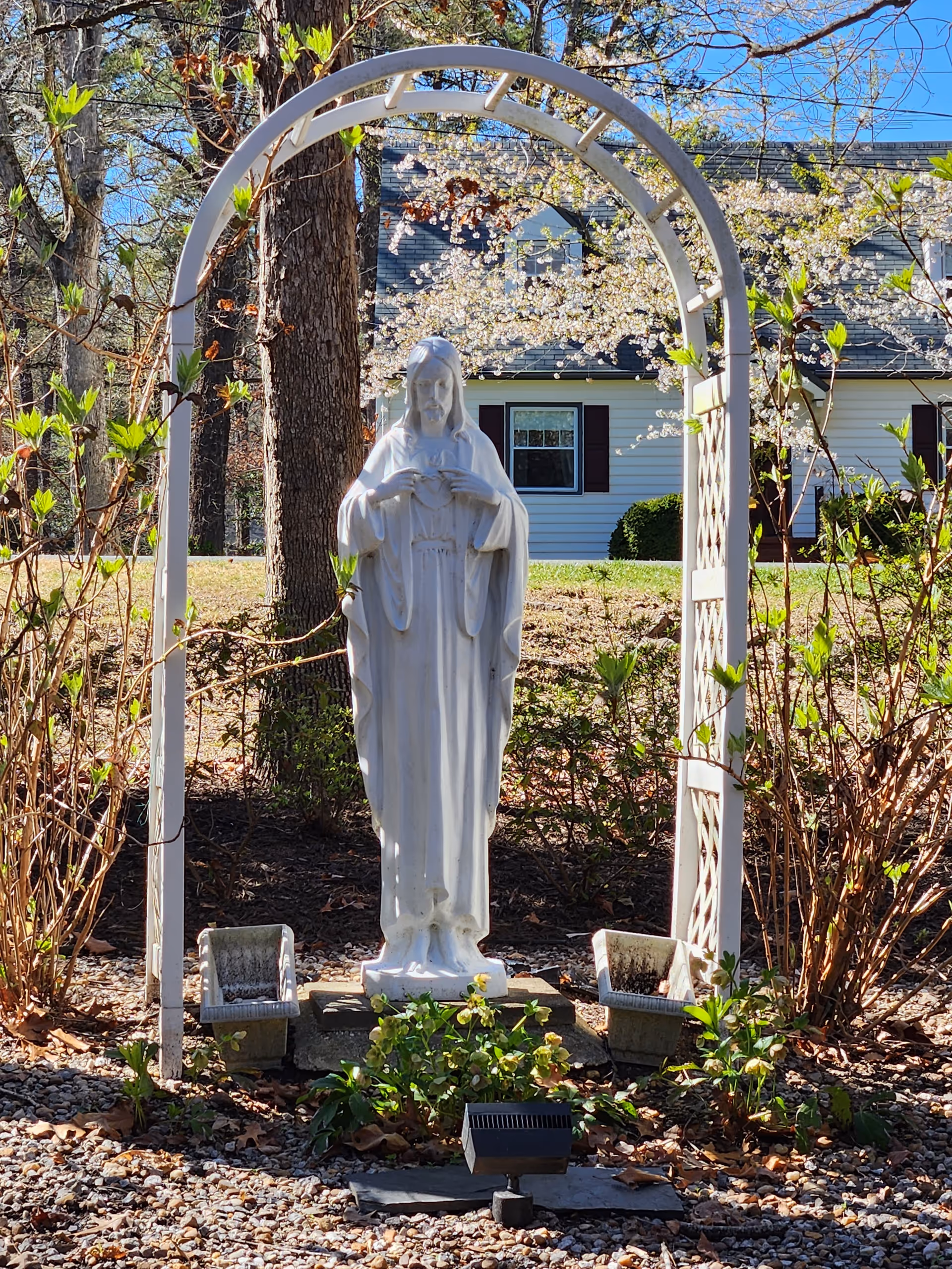 White statue of a religious figure under an arched trellis in a garden.
