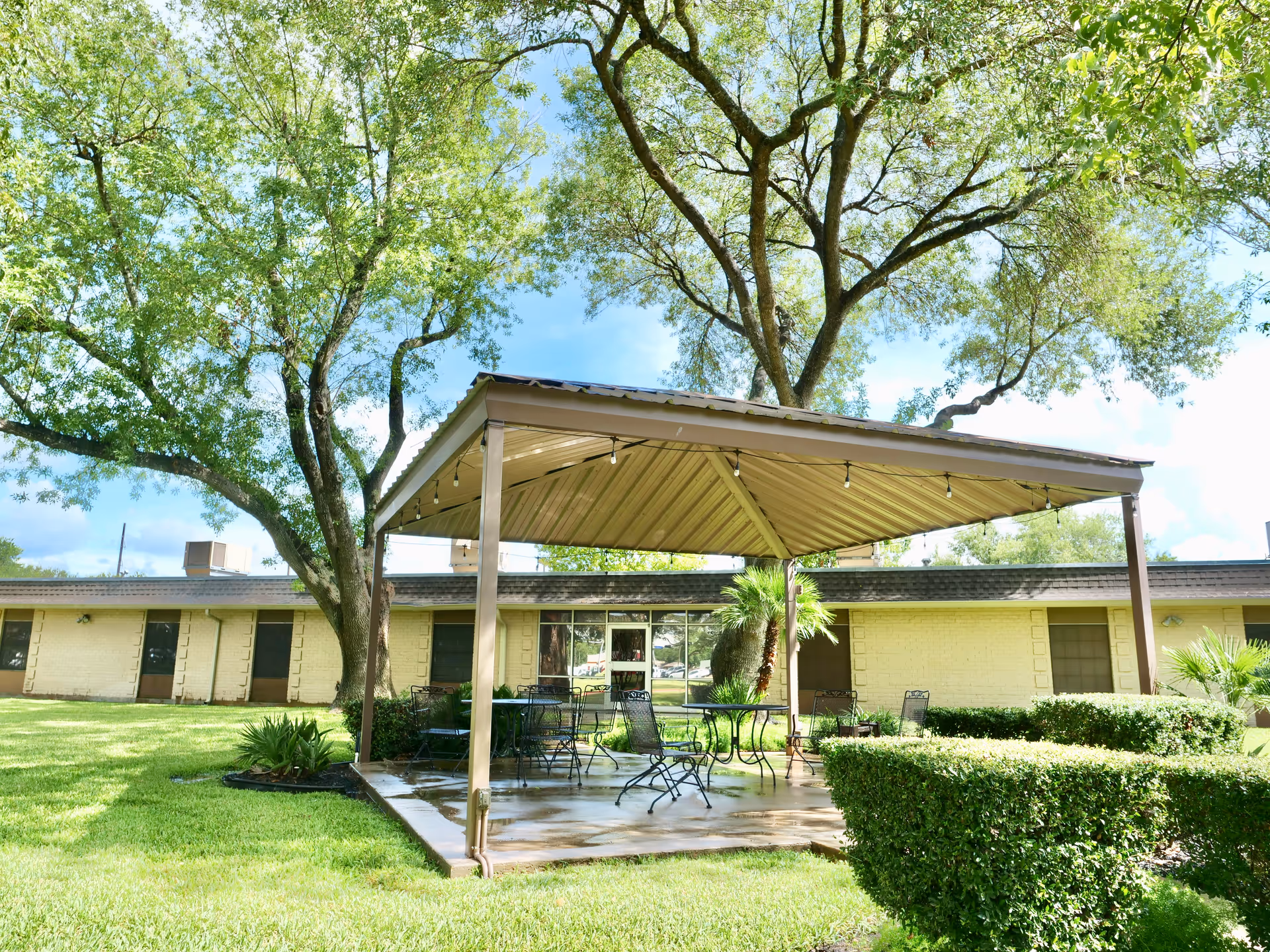 Outdoor patio area at Twin Pines Nursing & Rehab Center with a metal roof canopy, several metal tables and chairs, surrounded by green grass, trees, and trimmed bushes under a clear blue sky.