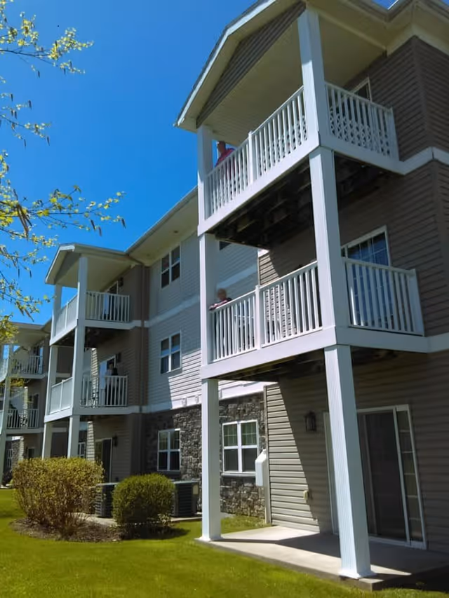 Exterior view of a multi-story residential building with balconies on each floor. The building has beige and brown siding with stone accents on the lower level. There are bushes and a well-maintained lawn in front of the building under a clear blue sky.