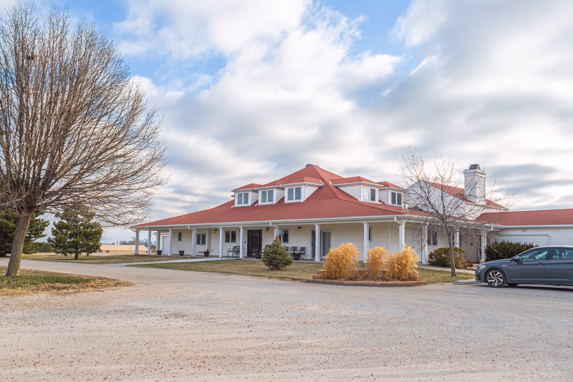 Senior living facility exterior: white siding building with red roof, driveway, car, and landscaping