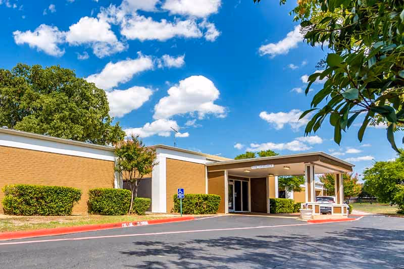 Front entrance of a one-story brick health and rehabilitation center with a covered drive-up canopy, parking area, and landscaping under a blue sky.