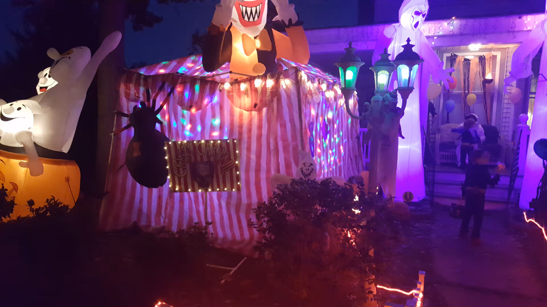 Outdoor Halloween display with striped tent, inflatable ghosts, and colorful lights around a residence.