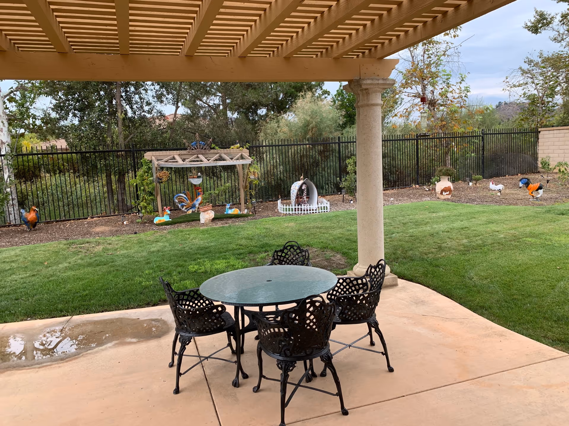 Outdoor patio area with a round glass table and four black metal chairs under a wooden pergola. Beyond the patio is a grassy lawn with decorative garden ornaments shaped like roosters and chickens, surrounded by a black metal fence and trees in the background.