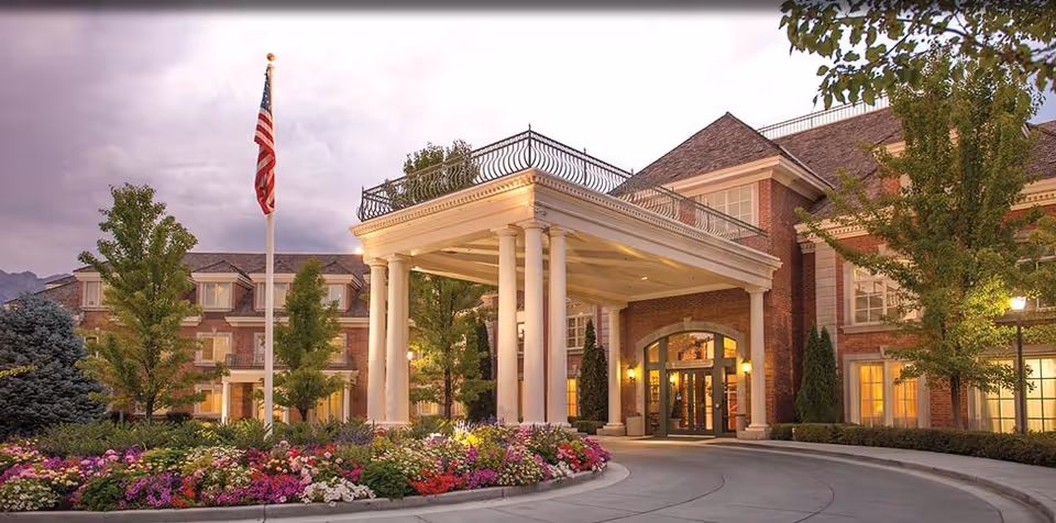 Brick senior living center with a portico entrance, columns, flag, and landscaped flower beds.