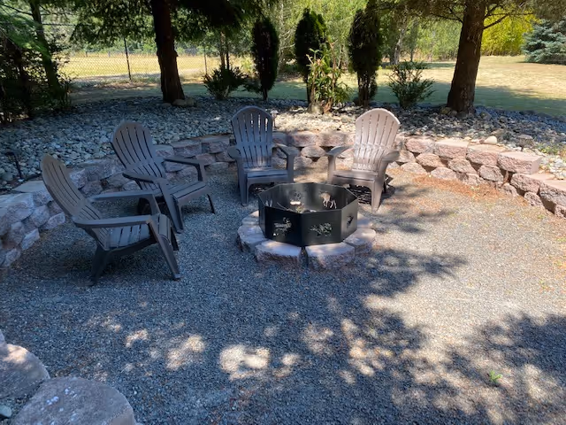 Outdoor fire pit surrounded by six gray Adirondack chairs in a gravel seating area.