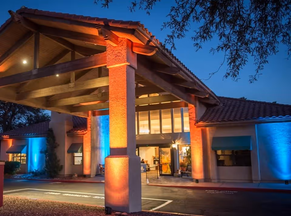 Exterior view of a senior living facility entrance at dusk with warm orange and blue lighting illuminating the building's columns and walls. The building has a tiled roof and large windows above the entrance.