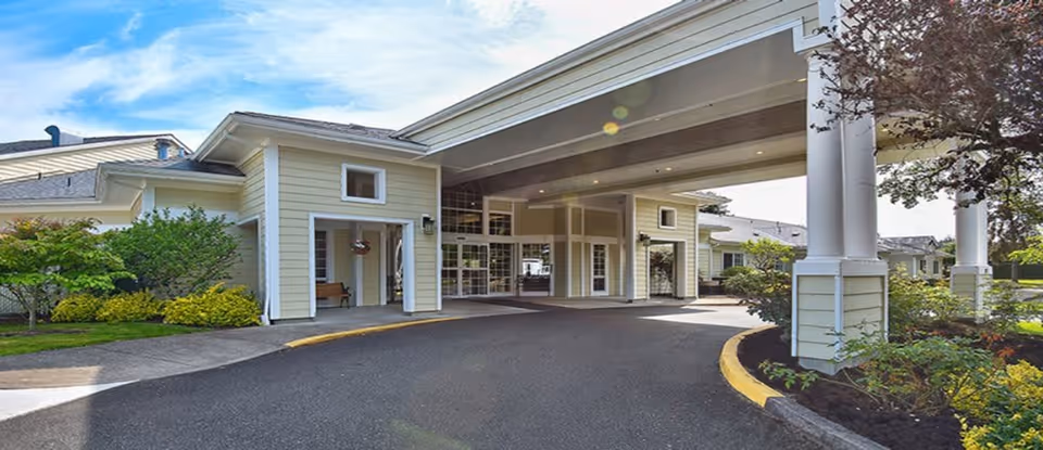 Exterior view of Deerfield Village Assisted Living facility showing the main entrance with a covered driveway, surrounded by landscaped greenery and trees under a partly cloudy sky.