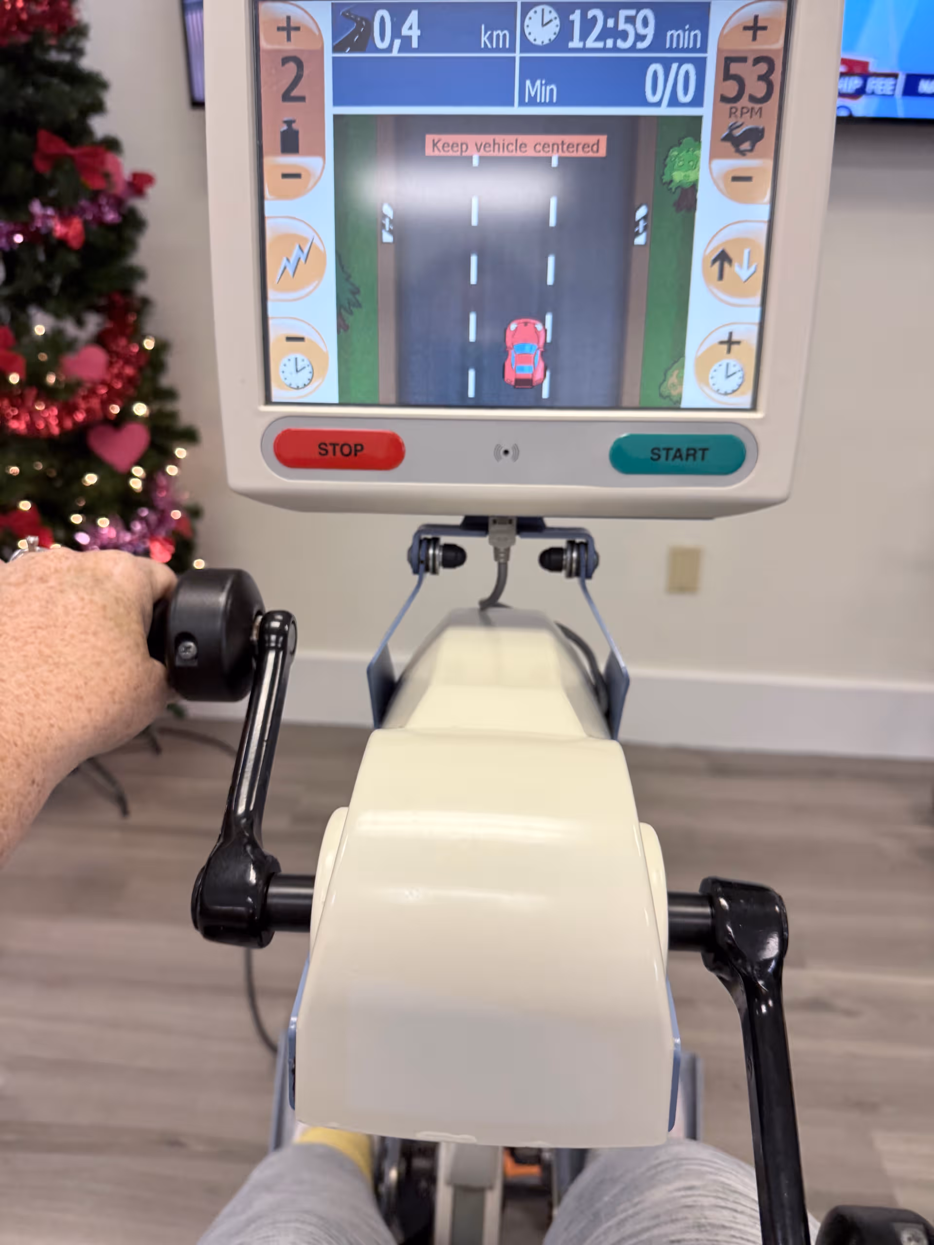Senior exercise bike with screen and hand on the handlebars, Christmas tree visible in background.