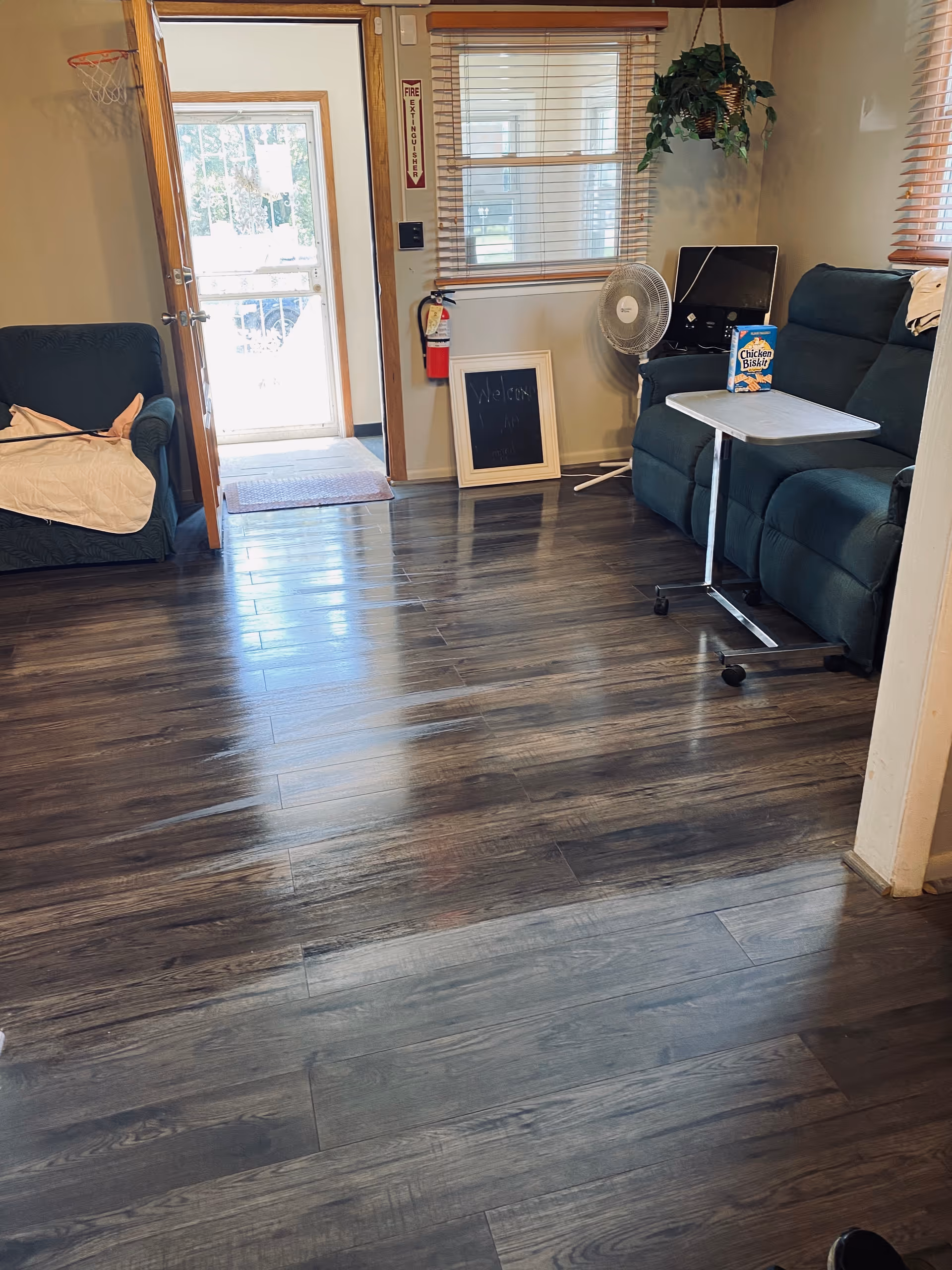 A living room with dark wood flooring, a dark green couch with a small rolling table holding a box of Chicken in a Biskit crackers, a standing fan, a window with blinds, a fire extinguisher mounted on the wall, an open door leading outside, and a small chalkboard on the floor near the window.