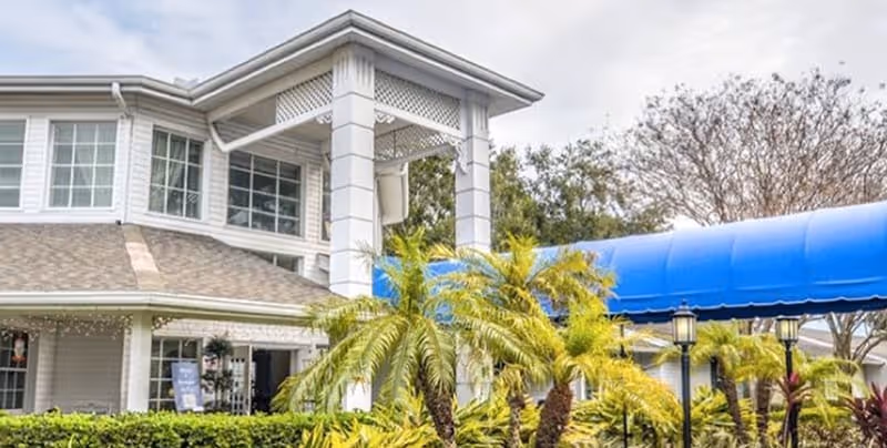 Exterior view of a senior living facility named The Pointe featuring a white building with large windows, a covered entrance with white pillars, a blue canopy, and landscaped greenery including palm trees and bushes.