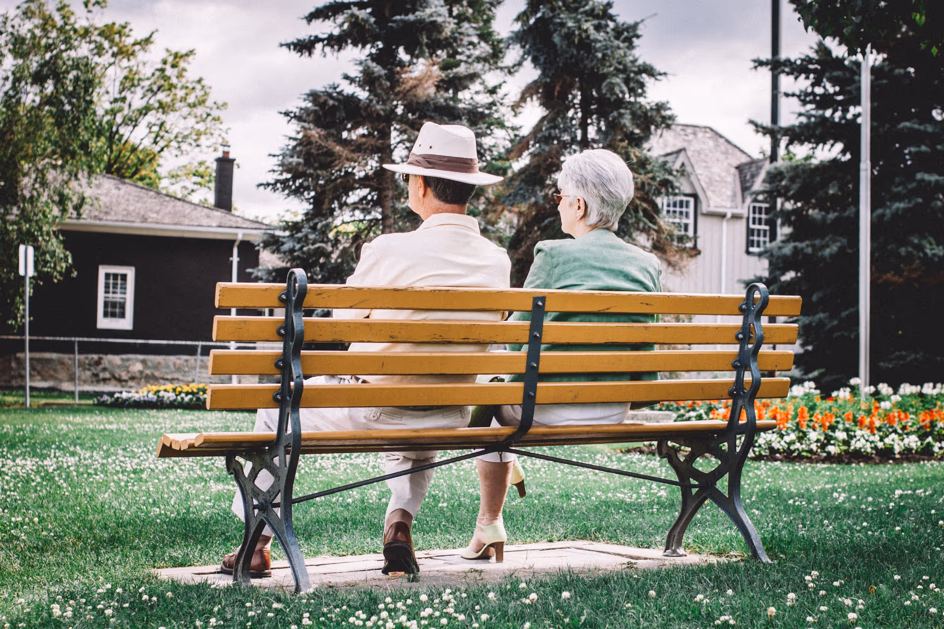 Two seniors sit on a wooden bench in a sunny outdoor park with flowers nearby today.