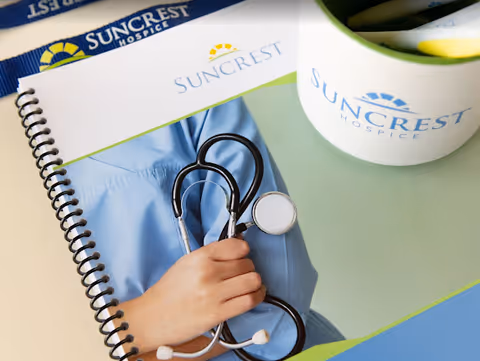 A hand holding a stethoscope rests on a Suncrest Hospice notebook.