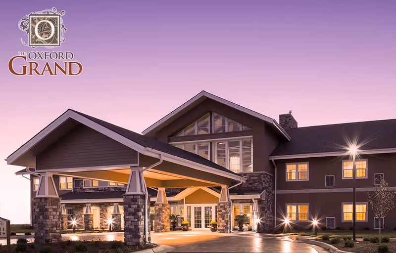 Exterior view of The Oxford Grand senior living facility at dusk, showing a large covered entrance with stone pillars, warm interior lights glowing through windows, and a clear sky with a purple-pink gradient.