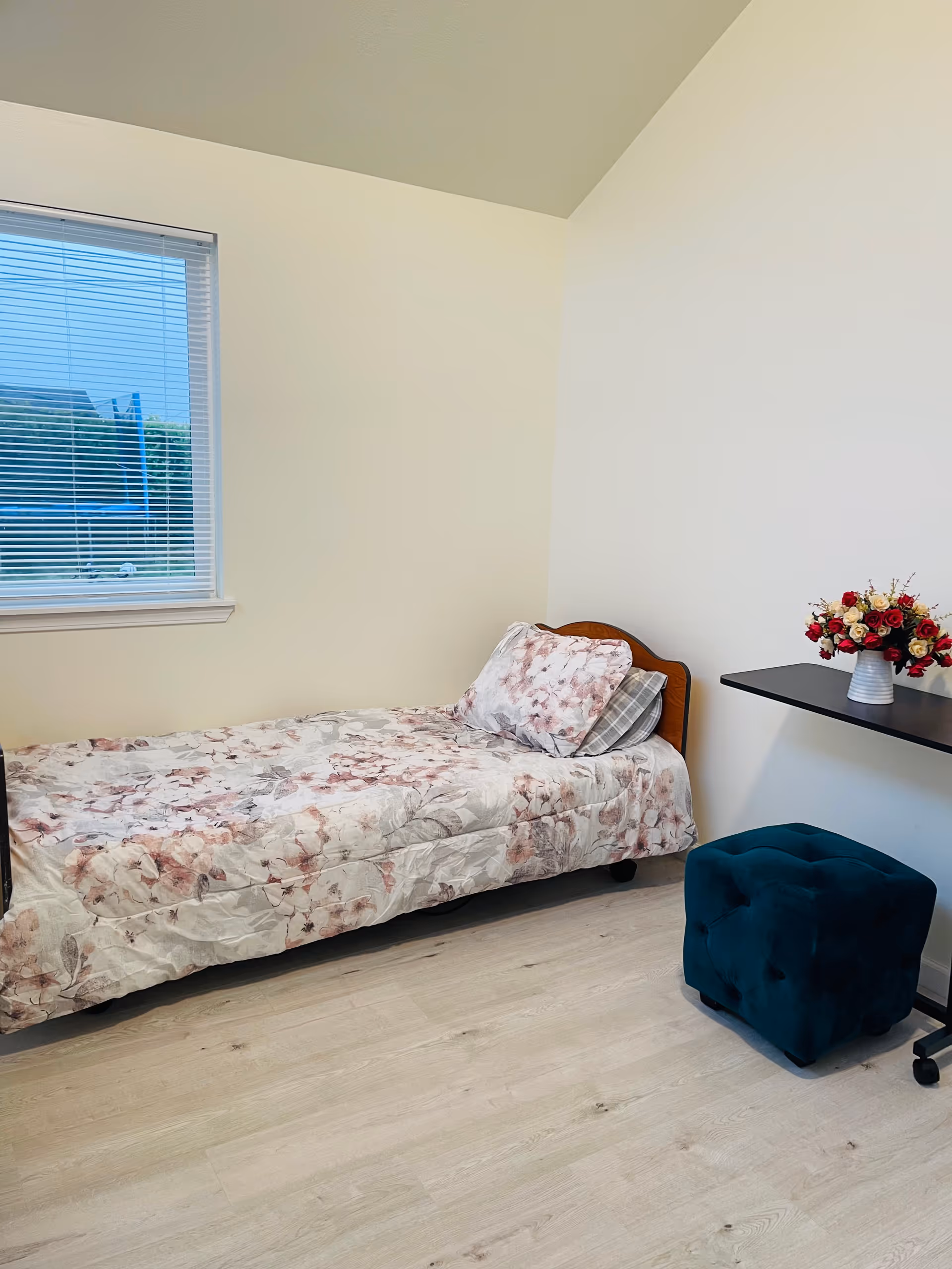 Single bedroom with floral bedspread, window, desk, and blue ottoman.