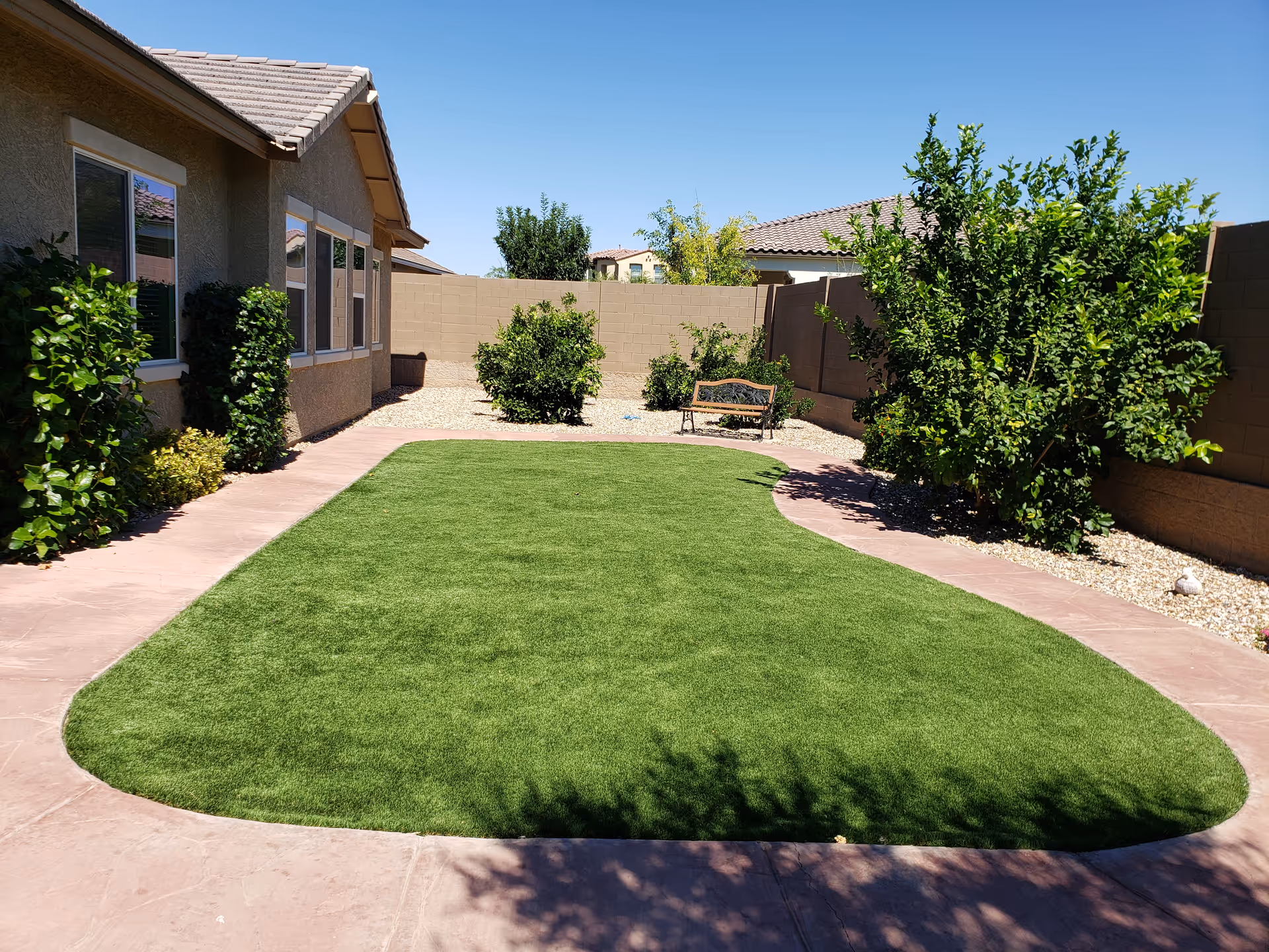 Outdoor courtyard with artificial lawn, curved pink pathways, shrubs, and a bench beside a house.