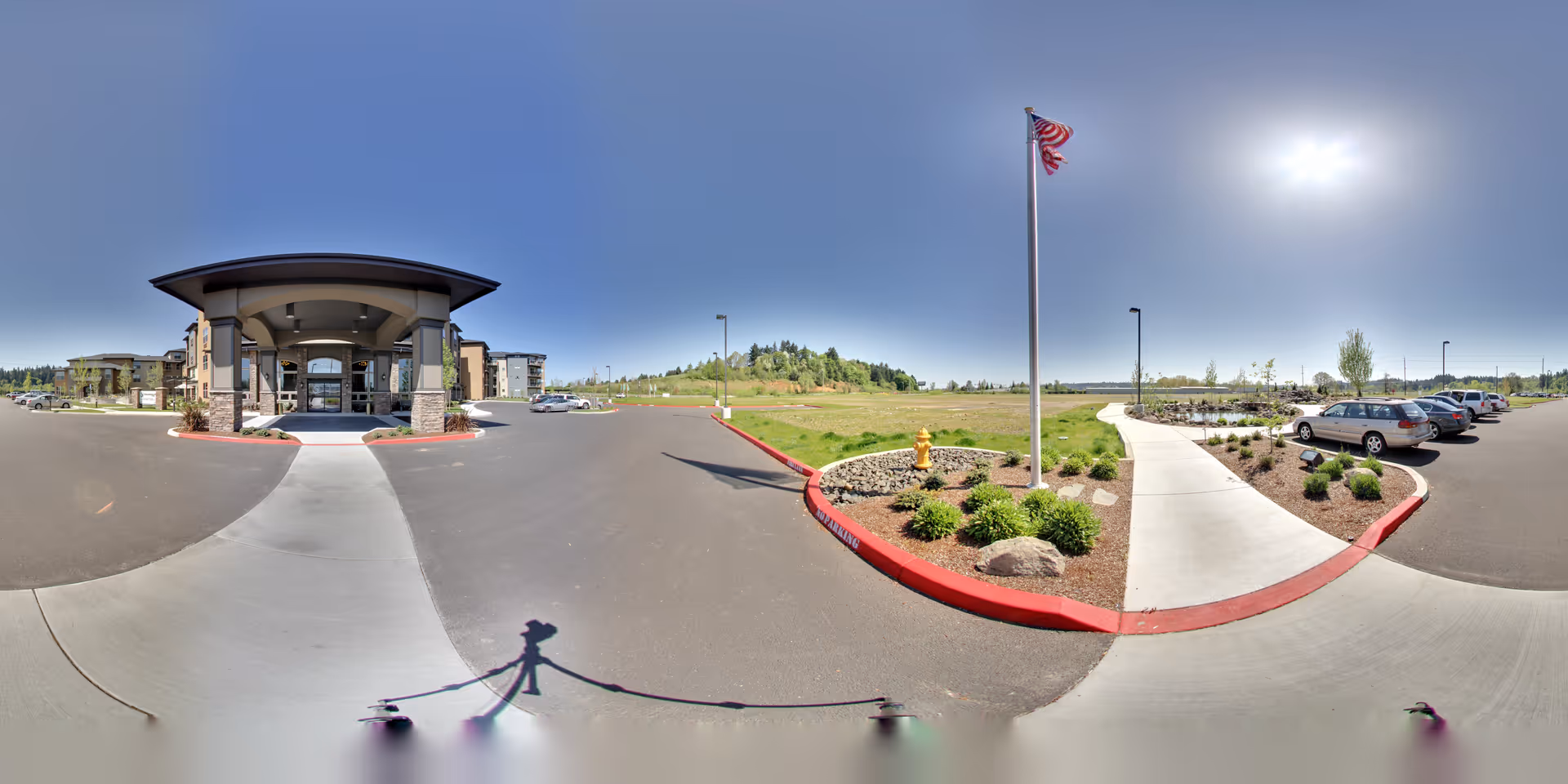 Wide panoramic view of the front entrance of a senior living facility named Bonaventure of Salem. The image shows a covered entryway with stone pillars, a parking lot with several cars, a landscaped area with bushes and a fire hydrant, a sidewalk, and an American flag on a flagpole under a clear blue sky with the sun shining brightly.