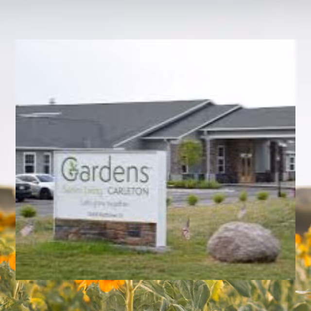 Exterior view of the Gardens of Carleton facility showing a large sign with the facility name in front of a single-story building with a gray roof and a covered entrance. There is a grassy area with a large rock and some small plants near the sign.