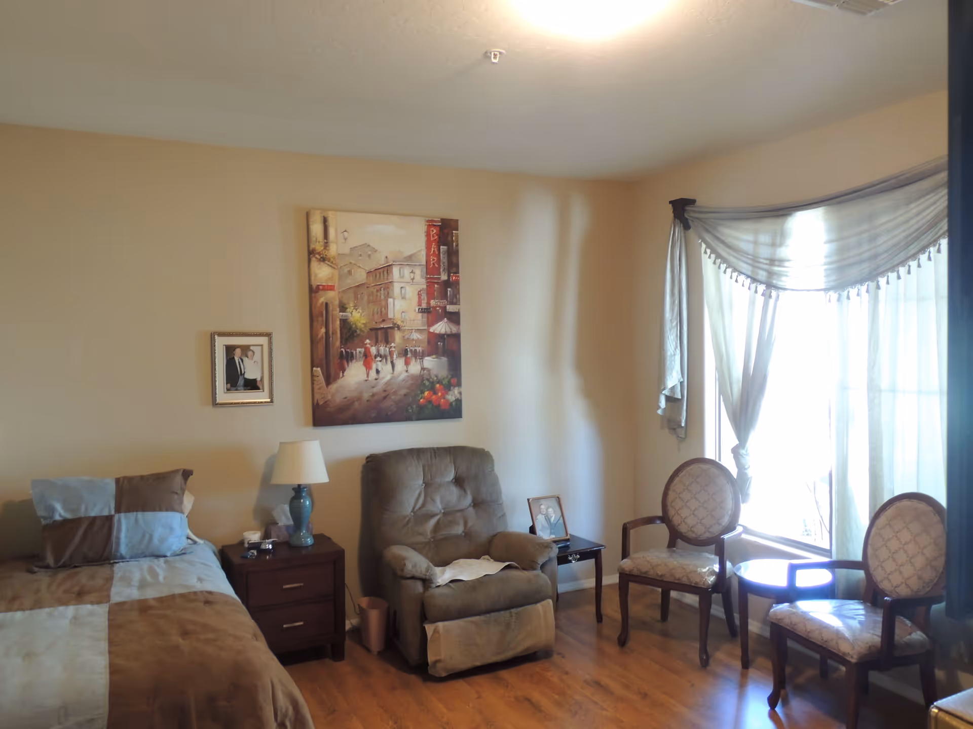 A cozy bedroom with a single bed covered in a brown and blue patchwork comforter, a wooden nightstand with a lamp and some items on it, a cushioned recliner chair, two upholstered chairs with a small round table between them near a window with sheer curtains, and two framed pictures hanging on the beige wall.