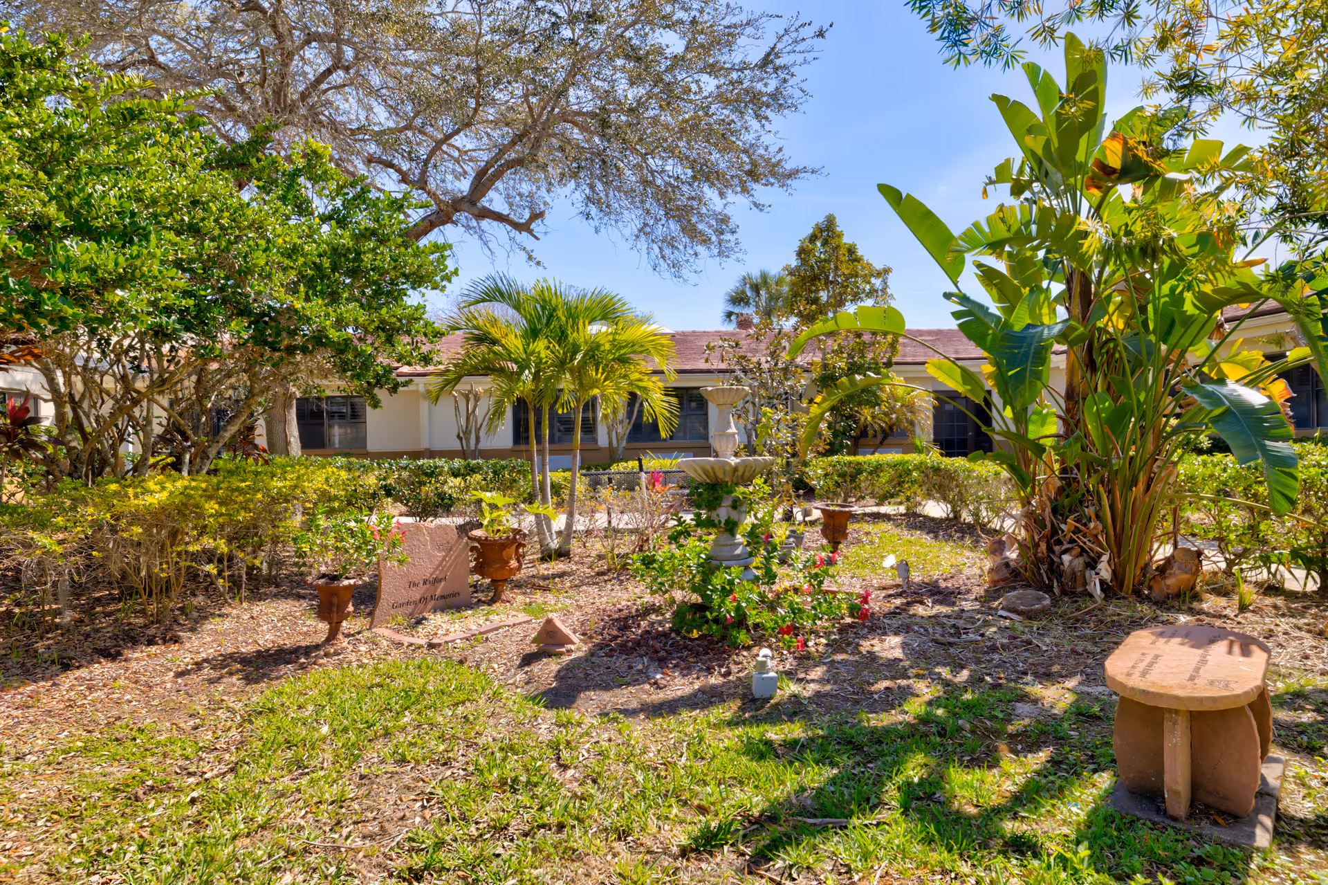 A sunny outdoor garden area at The Terrace of Kissimmee featuring various green plants, small trees, a stone birdbath, and a stone bench. The garden is surrounded by a building with windows and a red roof under a clear blue sky.