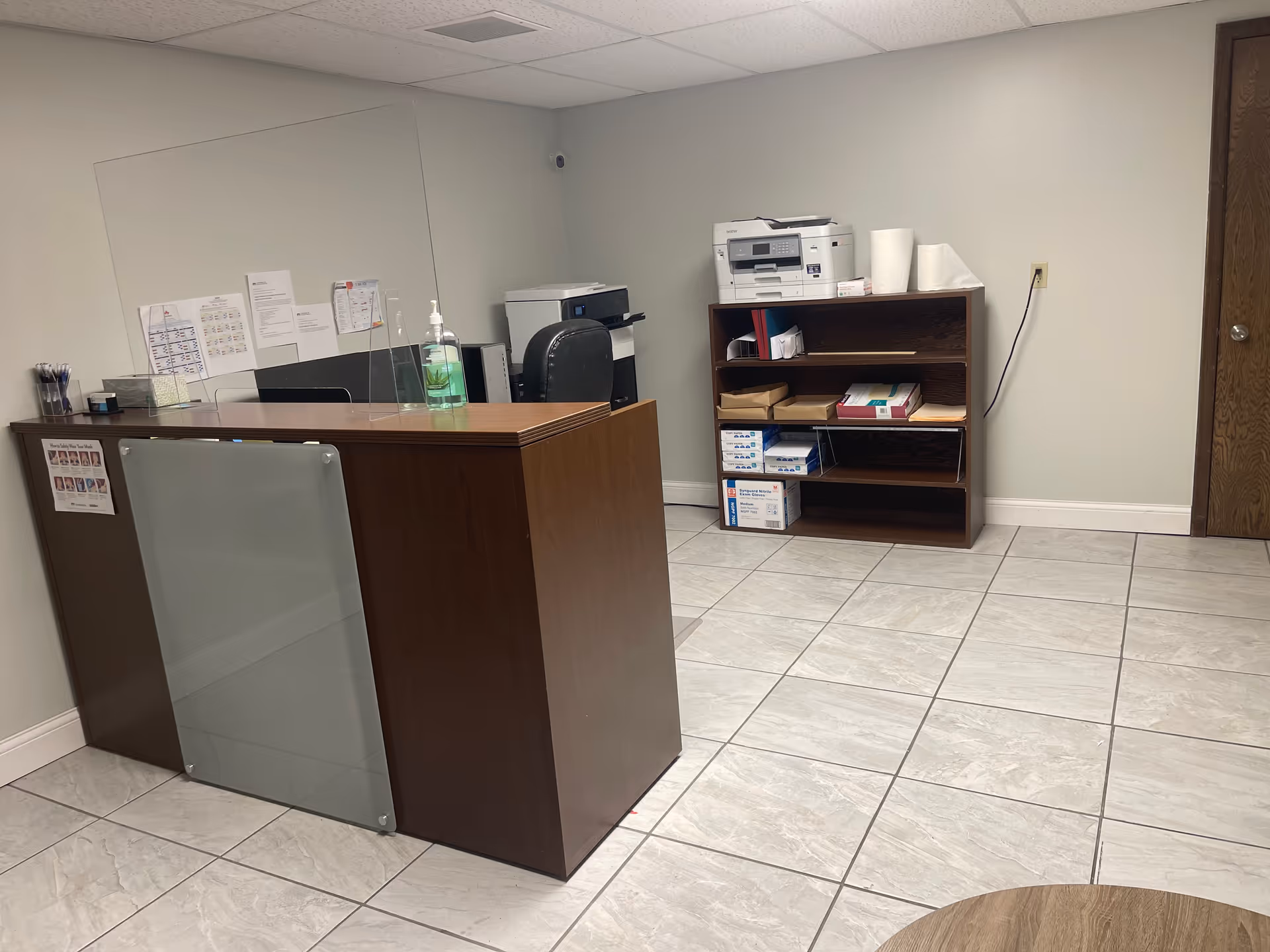 Reception area with dark wood desk, glass panel, copier, and shelving in a tiled lobby.