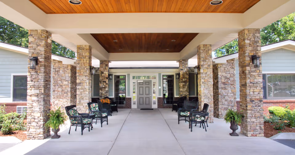 Covered entrance area of a senior living facility with stone pillars, black wicker chairs with floral cushions, potted plants, and a wooden ceiling. The entrance door is centered with windows on either side, and the building exterior features light gray siding and brick accents.