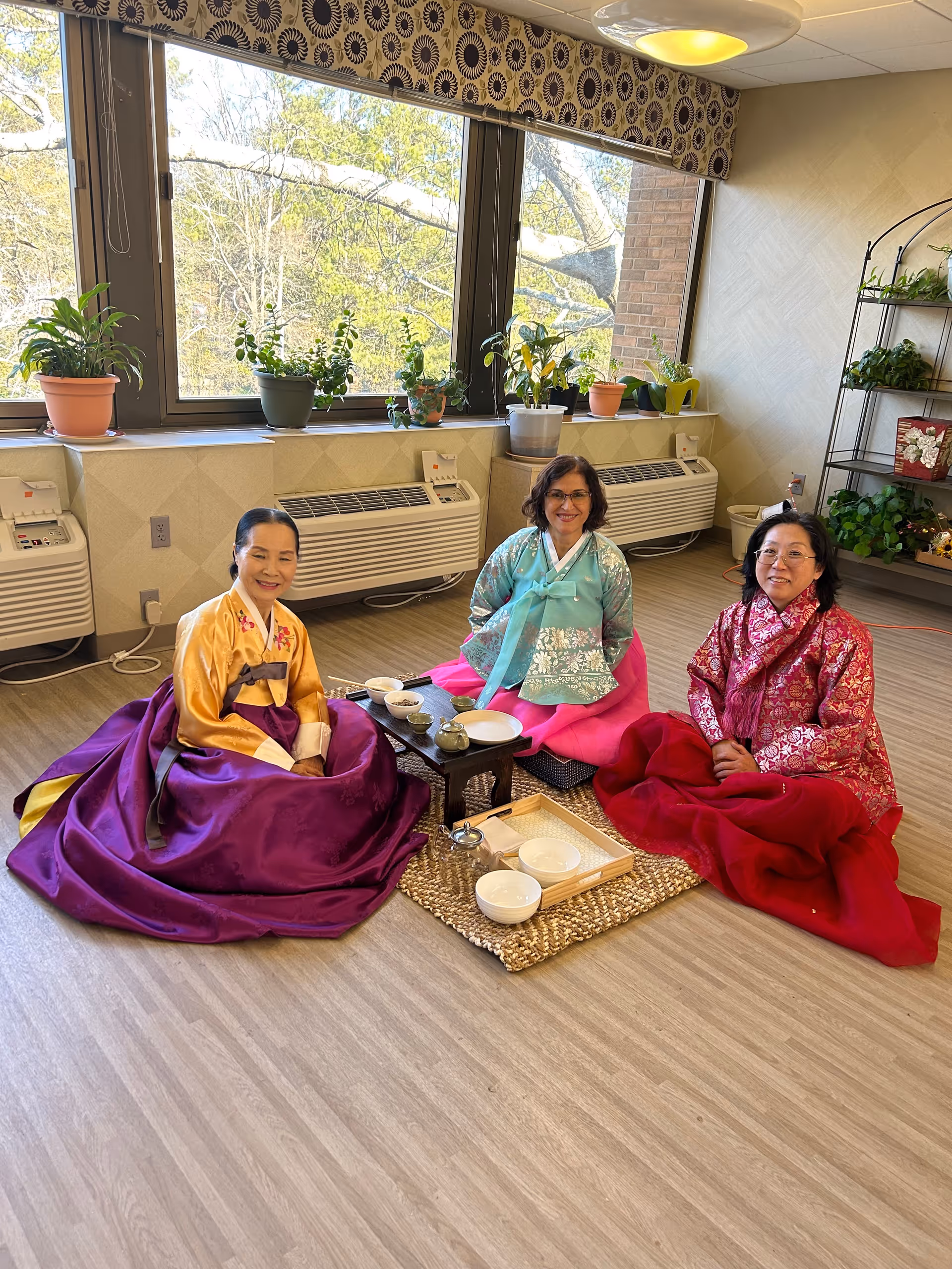 Three women dressed in traditional Korean hanbok sitting on the floor around a low table with tea sets in a bright room with large windows and several potted plants on the windowsill and a metal shelf.