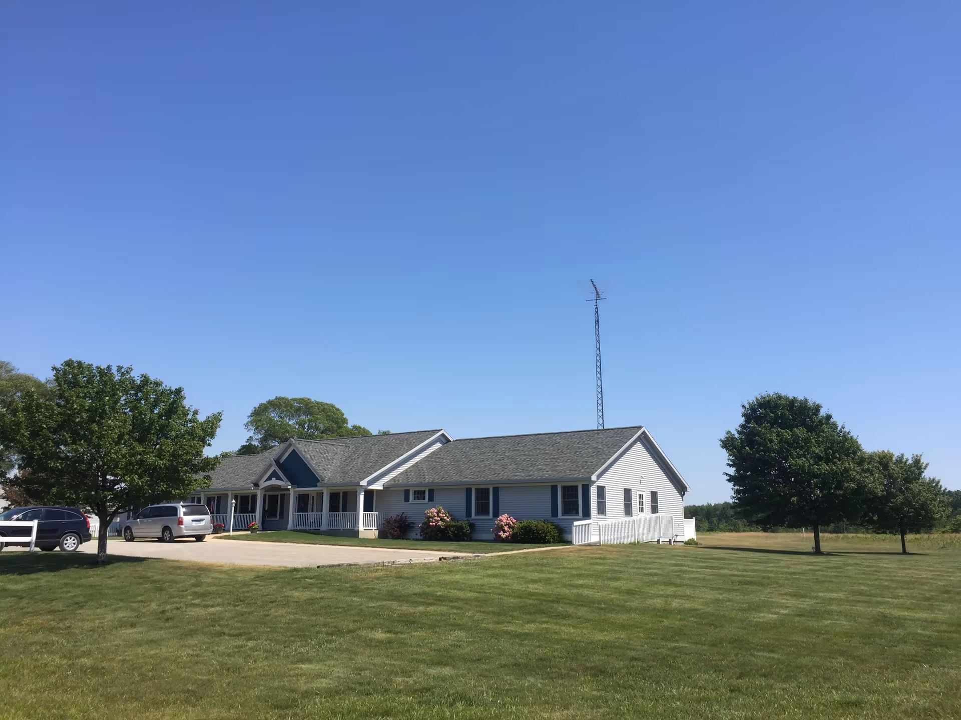 Single-story white building with a gray roof, surrounded by a large grassy lawn and a few trees under a clear blue sky. There are two cars parked near the building and a tall antenna behind it.