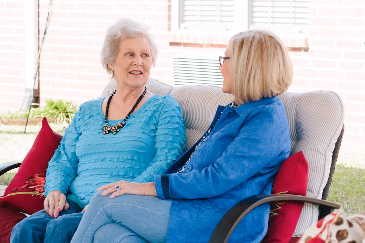 Two elderly women sitting and talking on a cushioned outdoor bench with red pillows, with a brick wall and grass visible in the background.