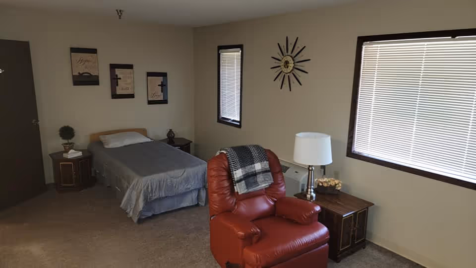 Cozy bedroom in a senior living facility featuring a bed, red recliner, and window blinds.