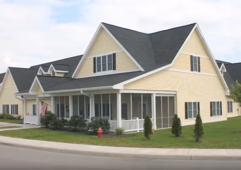 Exterior view of a large yellow building with a dark gray roof, featuring multiple gables and a screened porch. The building is surrounded by a well-maintained lawn with small shrubs and trees. An American flag is displayed near the porch, and a red fire hydrant is visible on the grass near the sidewalk.