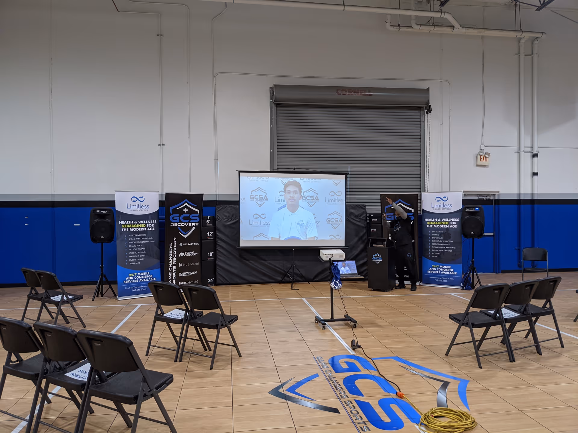 Indoor gymnasium setup with projector screen, banners, and empty folding chairs.