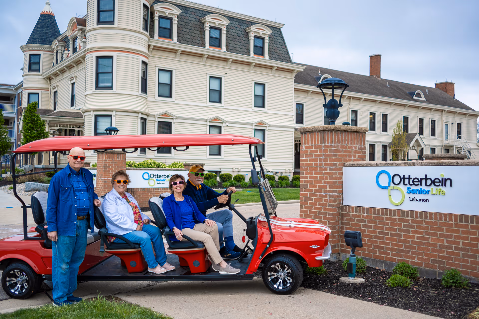 Five older adults pose on and beside a red golf cart in front of the Otterbein SeniorLife Lebanon building and entrance sign.