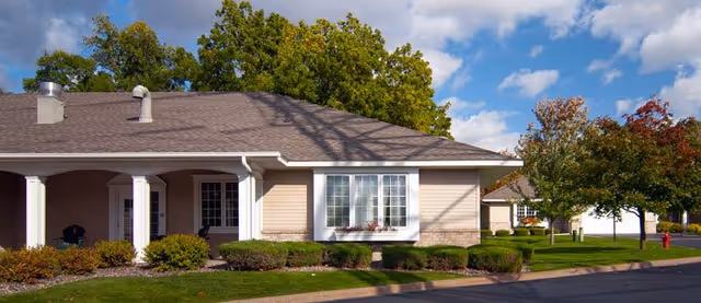 Front exterior of a single-story beige residential building with a covered porch, bay window, trimmed shrubs, and a tree-lined lawn under a blue sky.