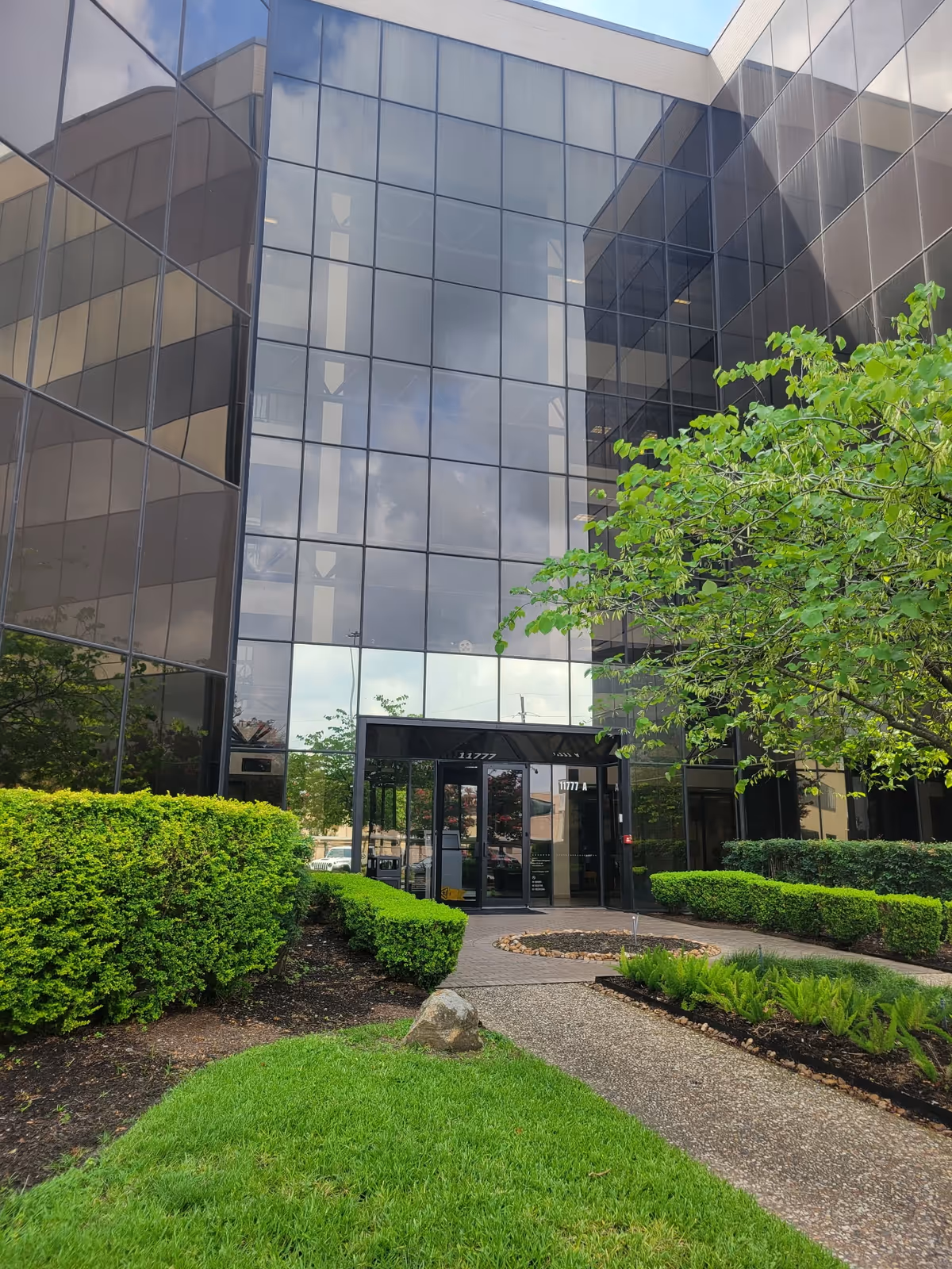 Glass-front office building entrance with shrubs and a paved walkway.