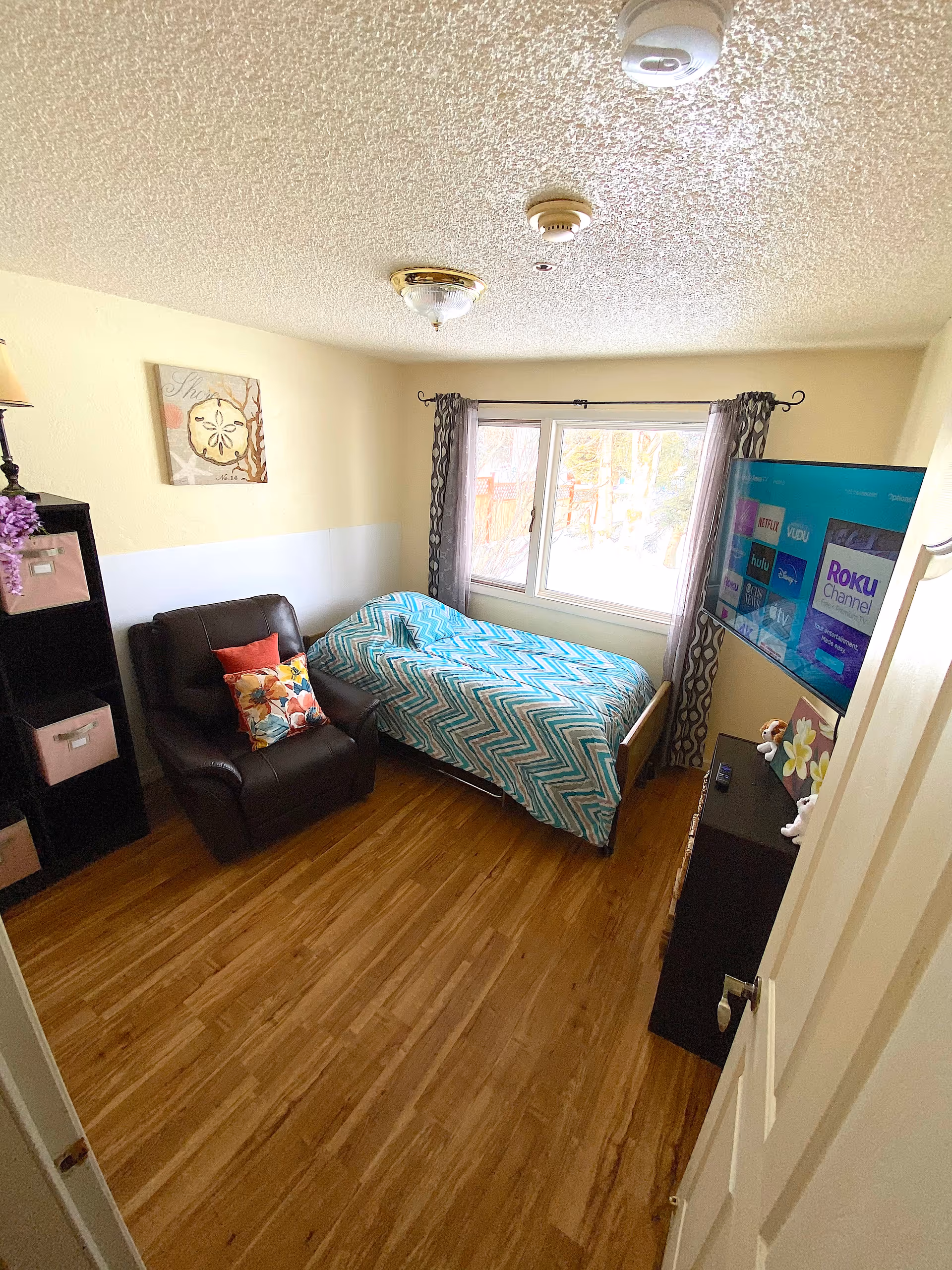 Cozy bedroom with bed, chair, dresser, TV, and window with curtains.