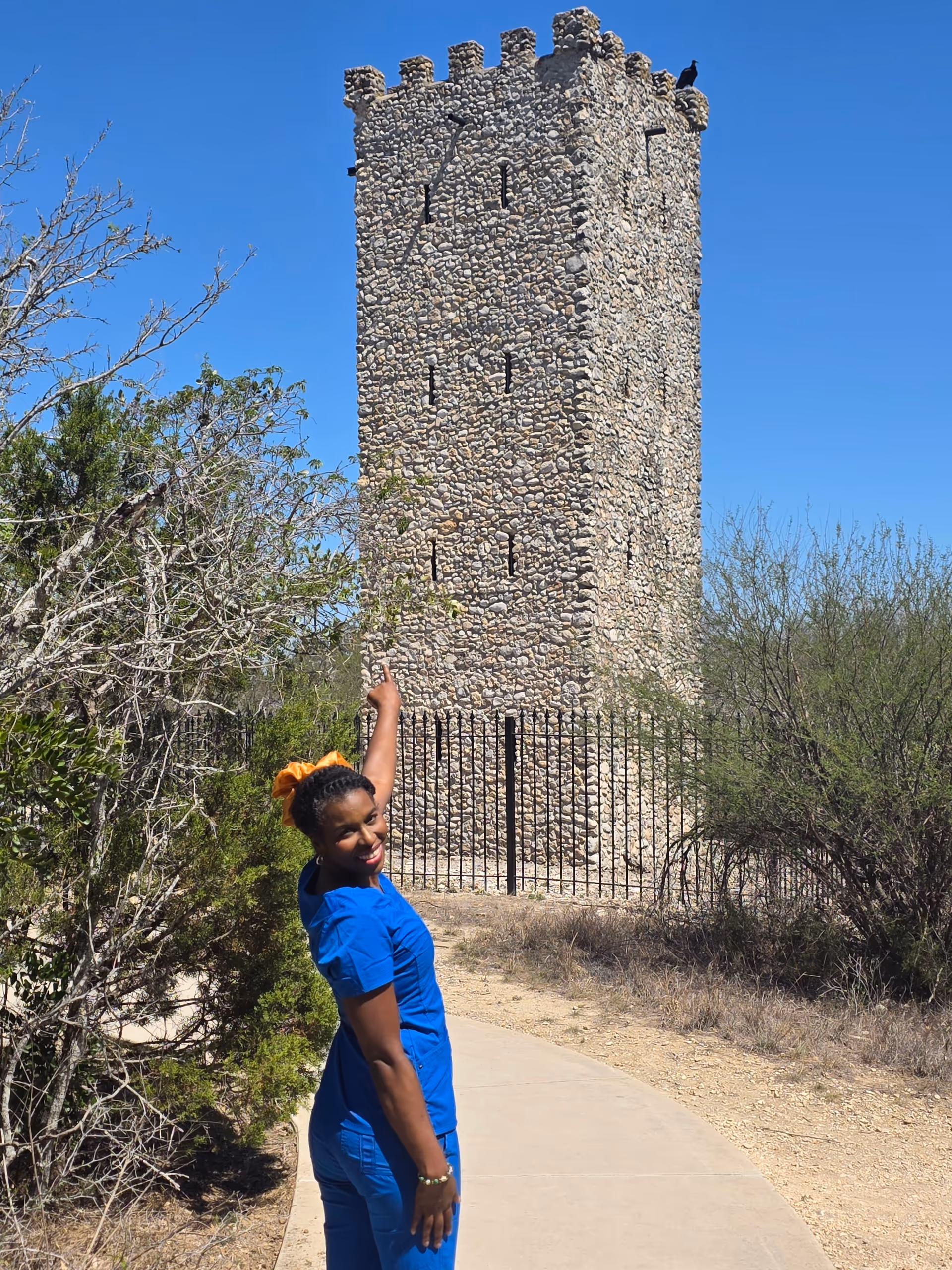 A woman in blue scrubs stands beside a tall stone tower outdoors along a walkway.