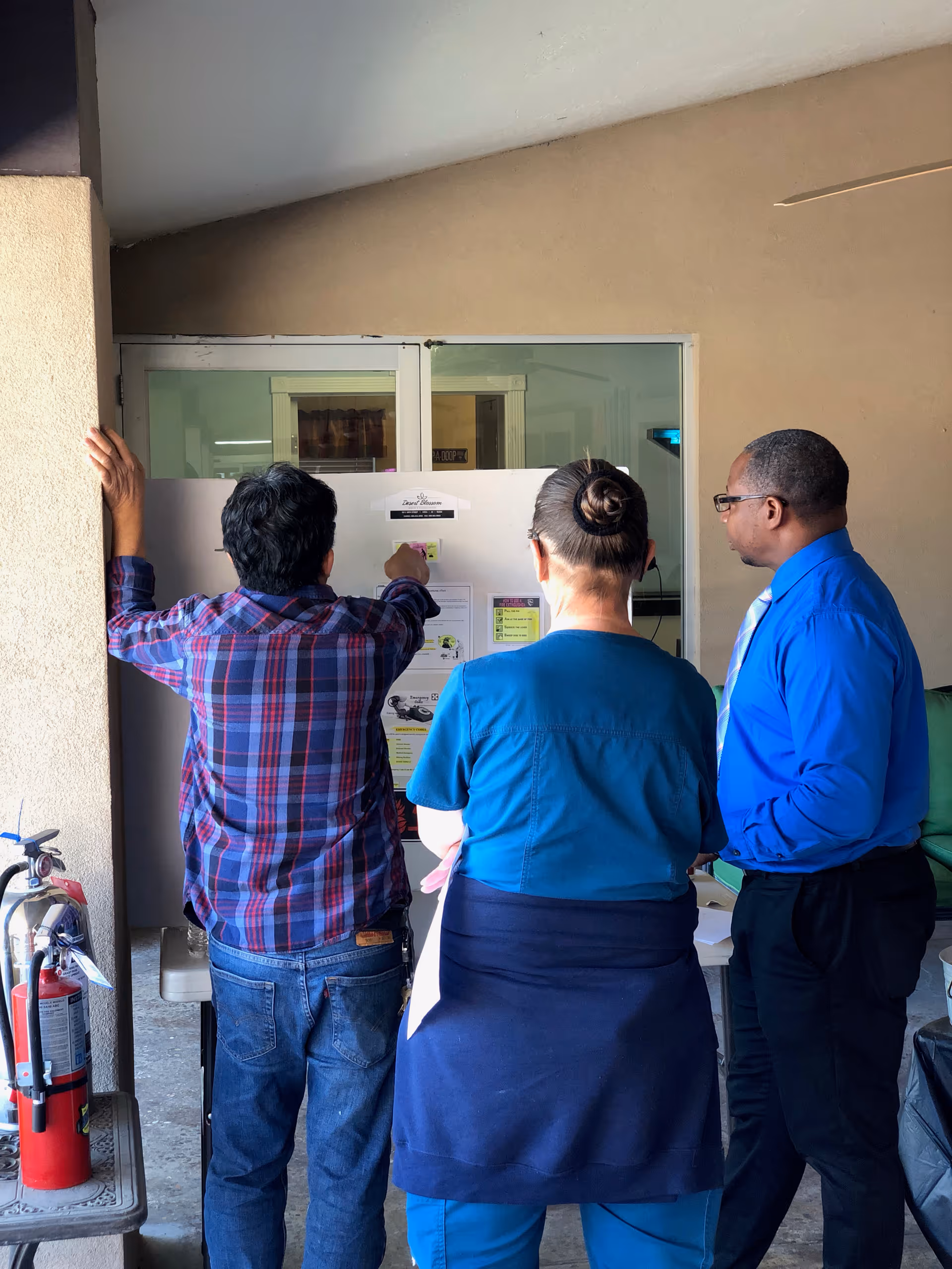Three people standing and looking at a bulletin board or information board mounted on a wall. One person is wearing a blue plaid shirt and jeans, another is in blue medical scrubs with a dark jacket tied around the waist, and the third person is wearing a blue dress shirt and black pants. A fire extinguisher is visible on the left side near a beige wall.
