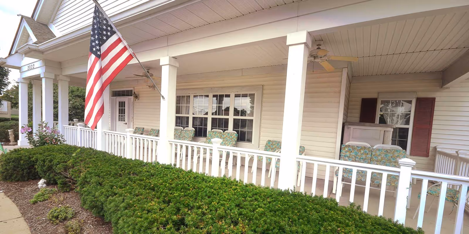 Front porch of a building with white railings and columns, several cushioned chairs with floral patterns, an American flag mounted on a pole, and green bushes along the walkway.
