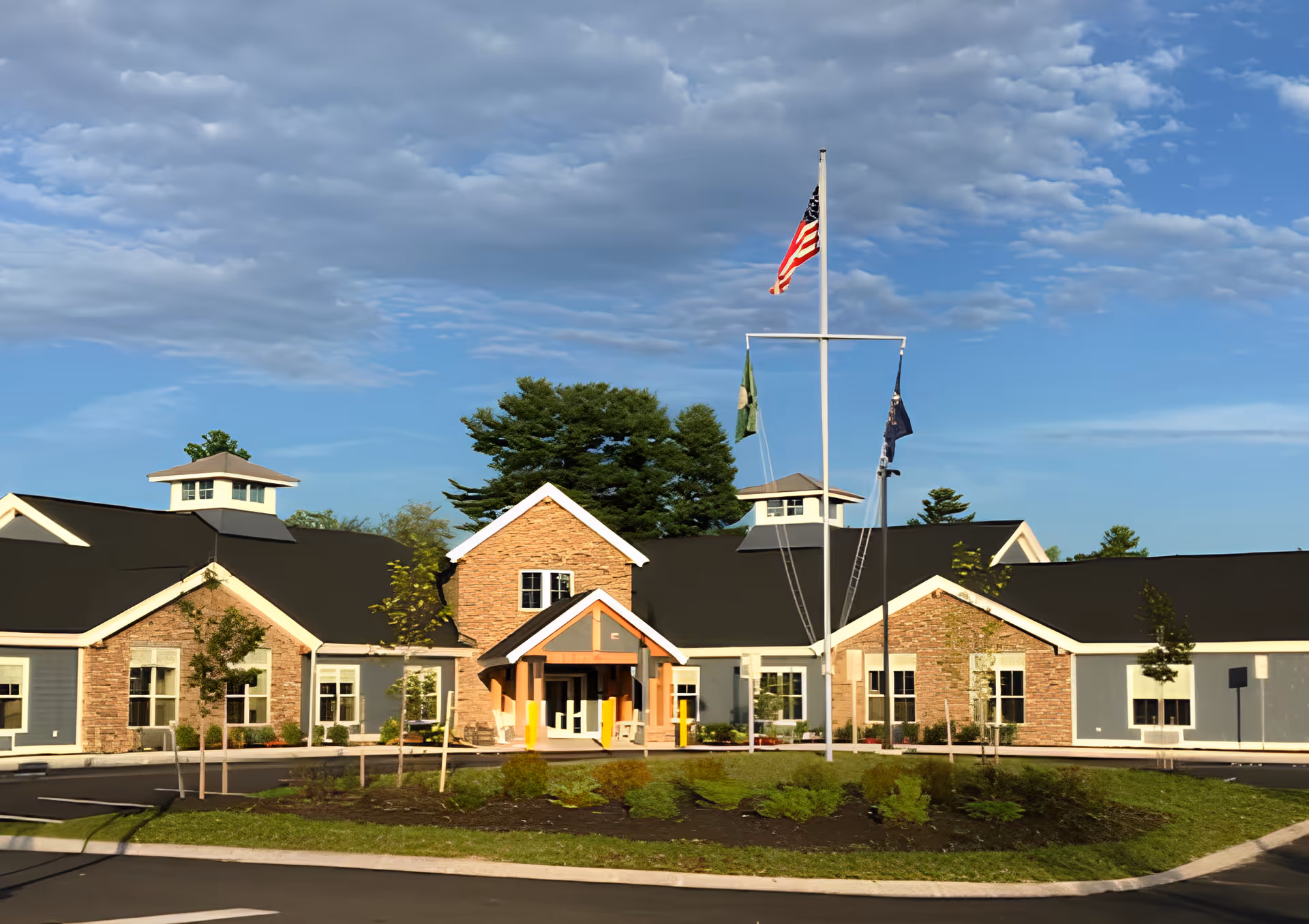 Front exterior view of a single-story senior living facility building with a combination of stone and blue siding, a covered entrance, and three flagpoles with flags including the American flag, under a partly cloudy blue sky.