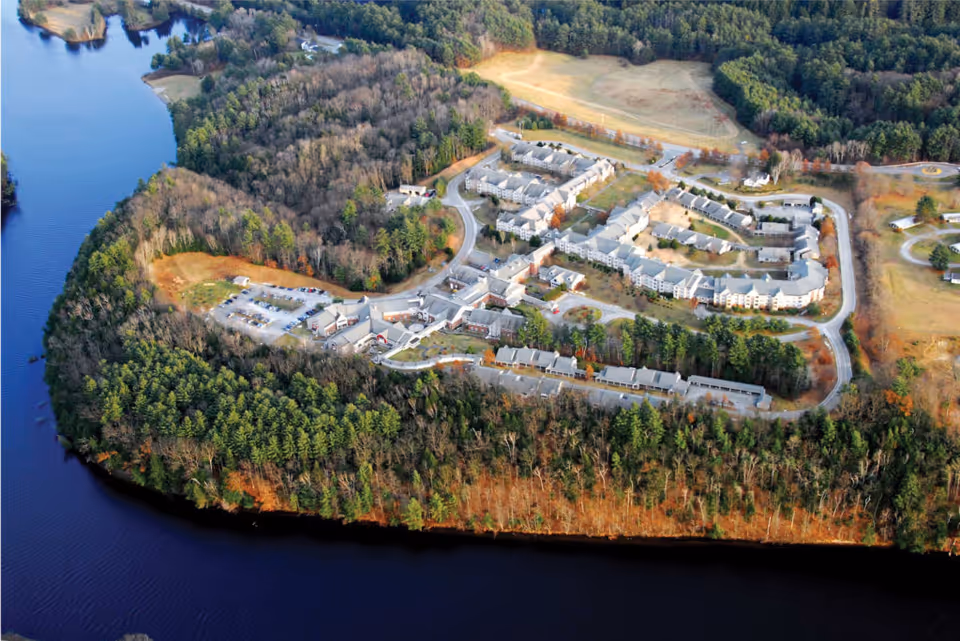 Aerial view of Kendal At Hanover senior living facility surrounded by dense trees and bordered by a large body of water. The campus includes multiple buildings, parking areas, and winding roads within a forested landscape.